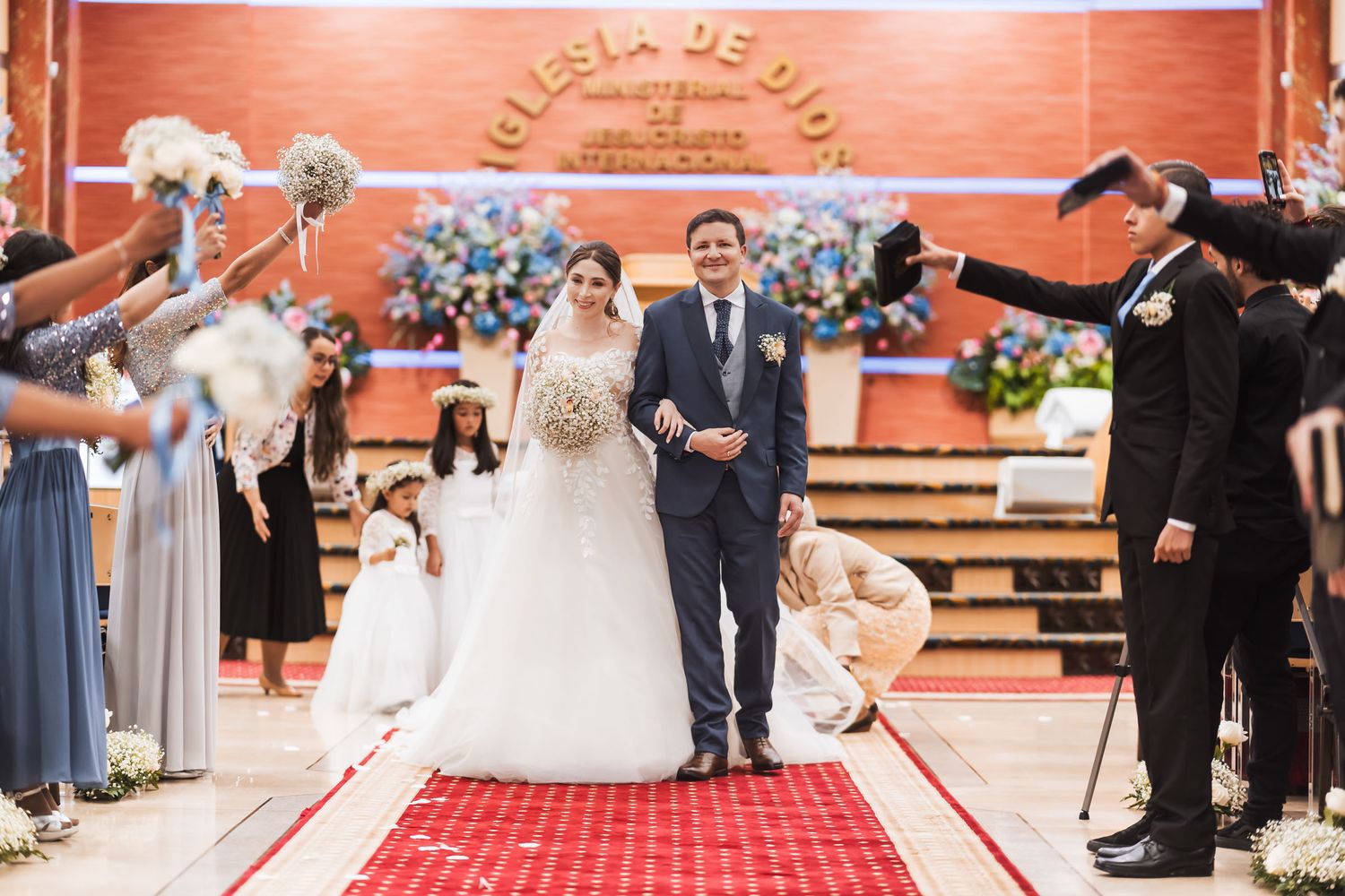 Salida nupcial: Novios caminan por pasillo rojo bajo arco de cortejo; niñas flores les siguen. Iglesia de Dios. Boda IDMJI.