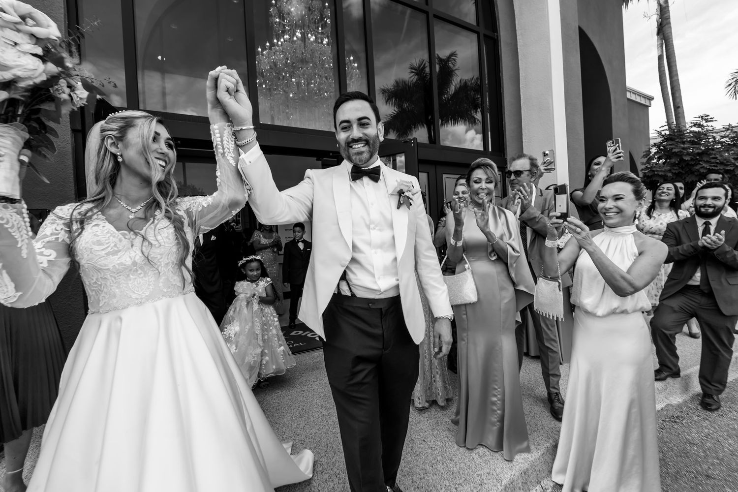 Foto B&W: Novios celebran con brazos alzados al salir de iglesia; invitados aplauden. Salida nupcial boda.