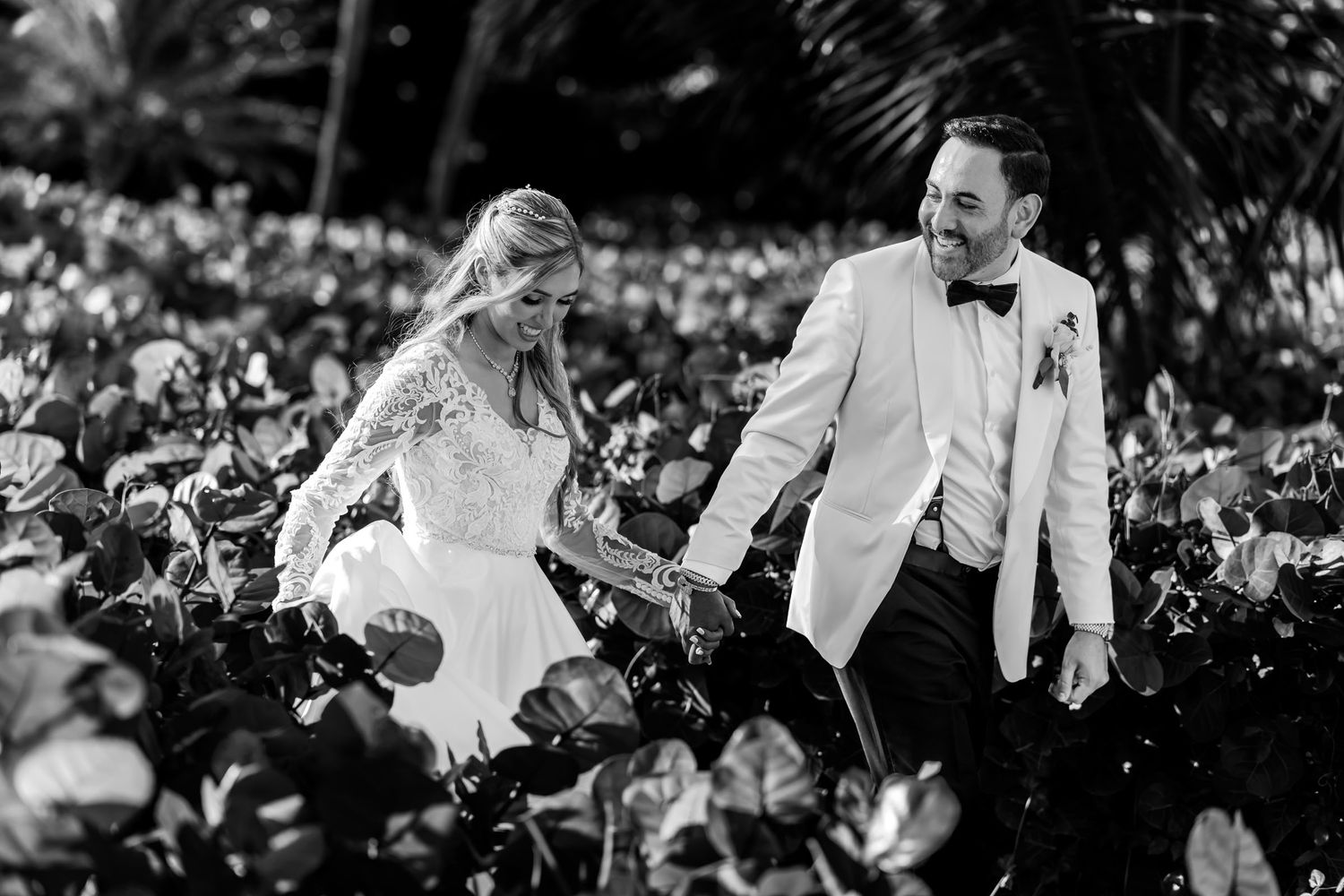 Foto B&W: Novios sonrientes tomados de la mano caminan entre vegetación tropical. Boda elegante.