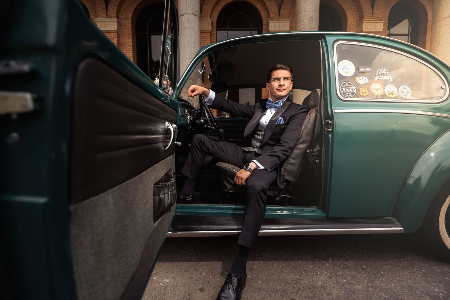 Novio elegante con esmoquin posa sentado al volante de coche clásico VW Escarabajo verde; puerta abierta. Boda.