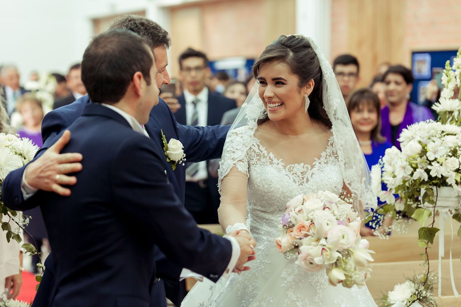 Novia radiiante con velo y ramo saluda a invitado o novio al llegar al altar; ceremonia boda.