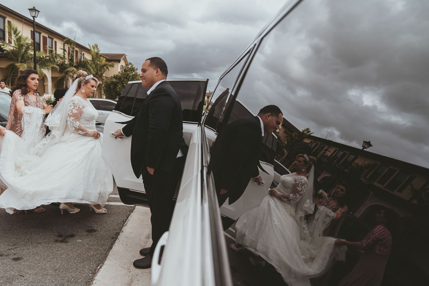 Novia y dama de honor bajan de limusina SUV blanca; chófer abre puerta, reflejo cielo nublado. Boda Miami.