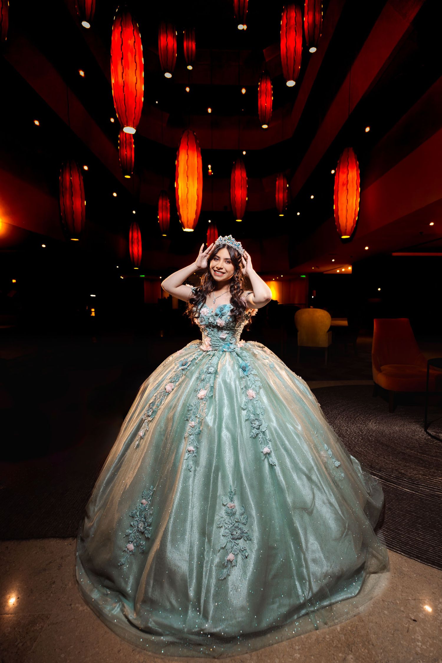 Quinceañera con vestido princesa aguamarina y tiara posa bajo linternas rojas colgantes en salón amplio.