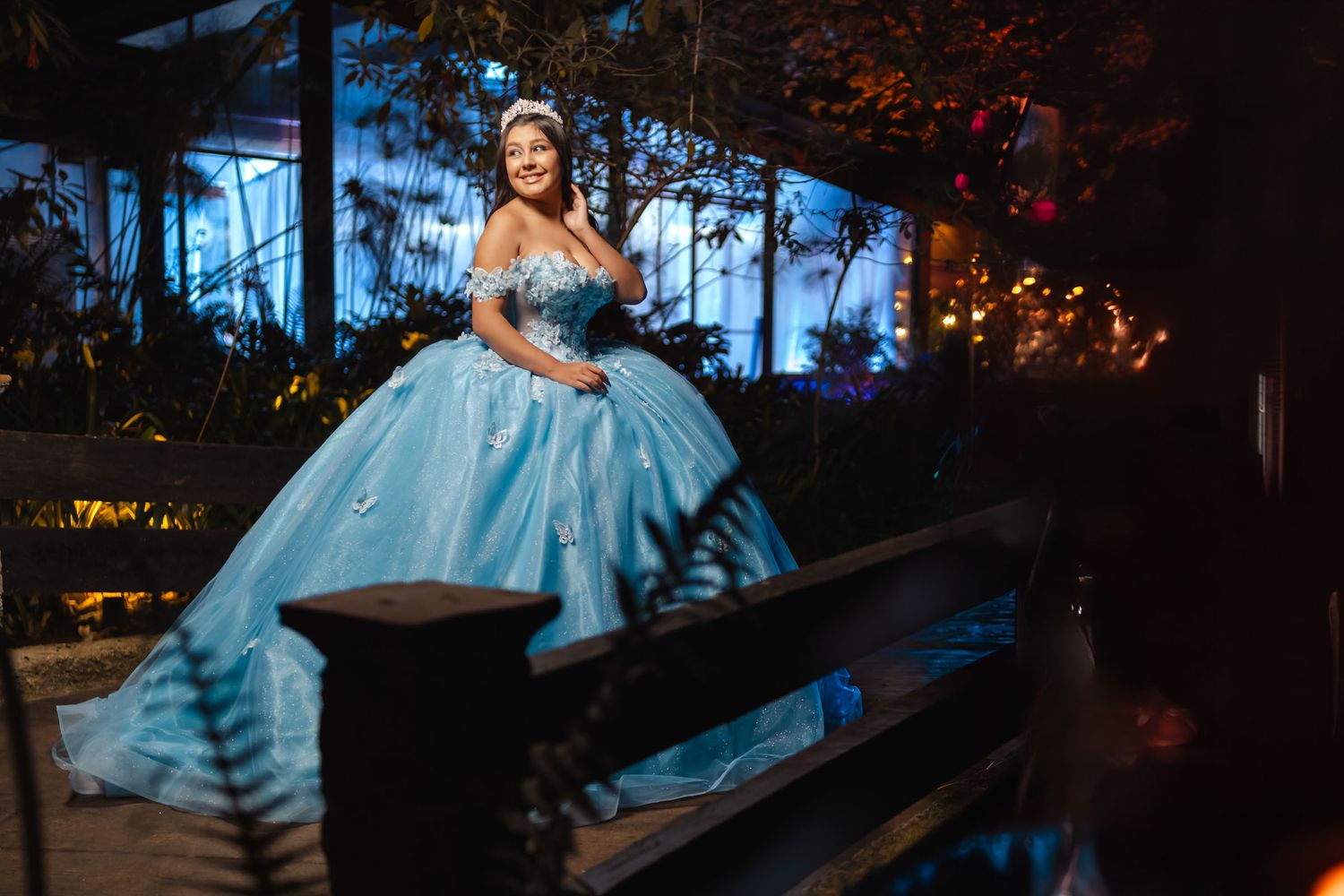 Quinceañera con vestido azul princesa y tiara sentada en puente de madera; fondo nocturno con luces.