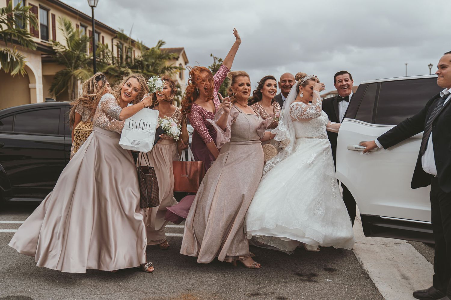 Novia y damas de honor celebran efusivamente al salir de vehículo blanco; chófer abre puerta. Día boda Miami.