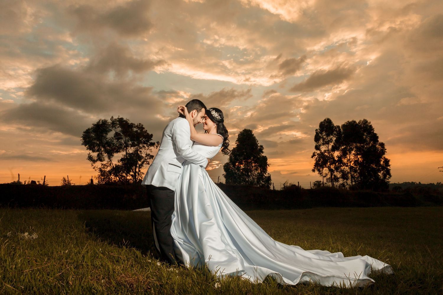 Novios se abrazan románticamente en campo de césped bajo espectacular cielo nublado al atardecer, durante hora dorada. Boda exterior.