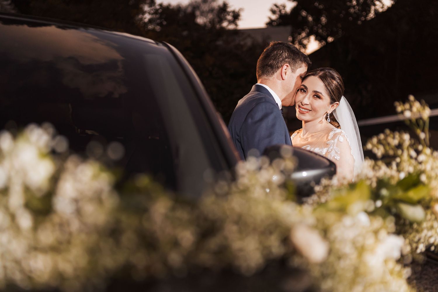 Novio besa mejilla de novia sonriente junto a coche oscuro decorado con flores blancas; foto boda nocturna romántica.