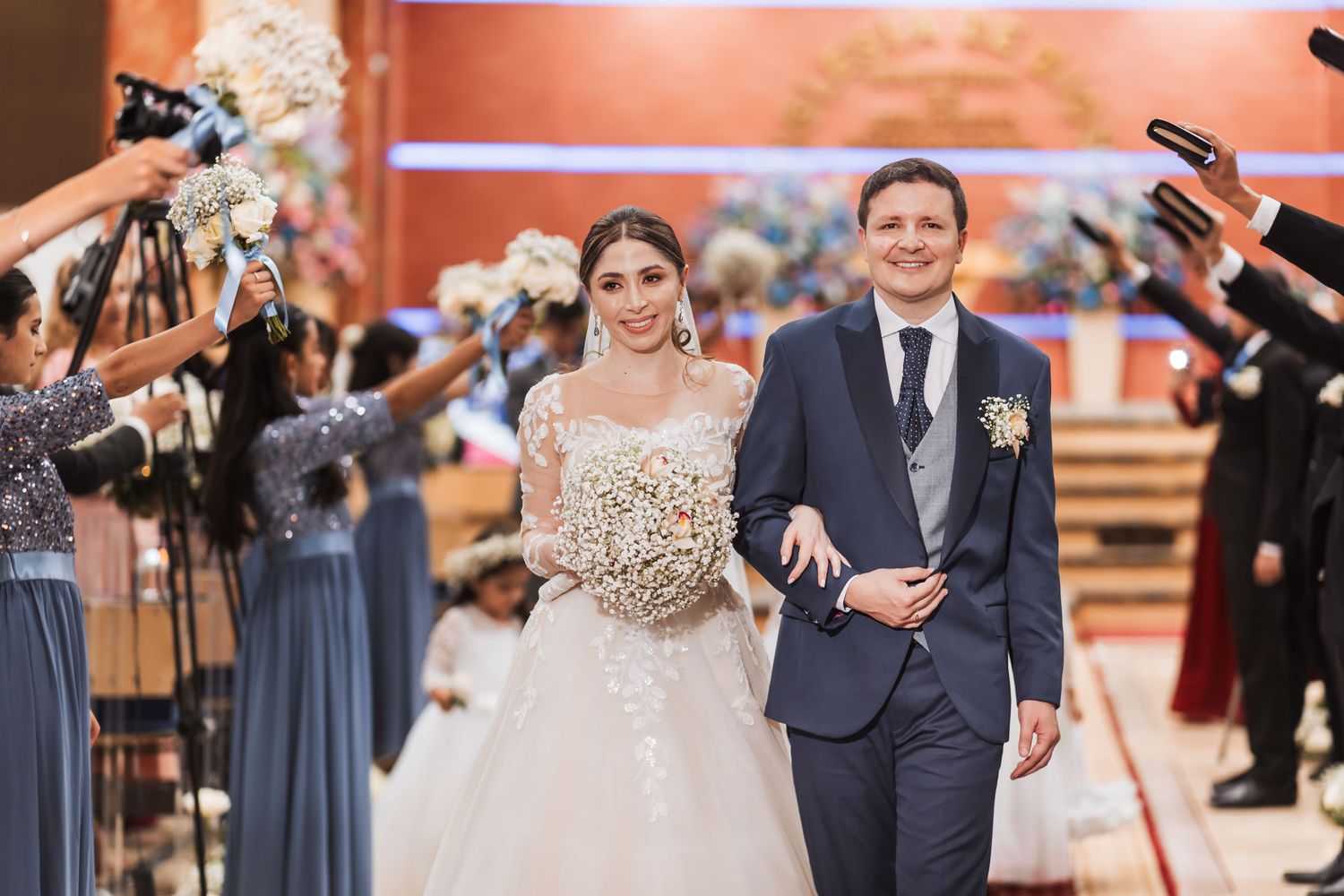Novios felices caminan bajo arco de cortejo en salida nupcial; novia ramo grande, novio traje azul. Iglesia. Boda IDMJI.