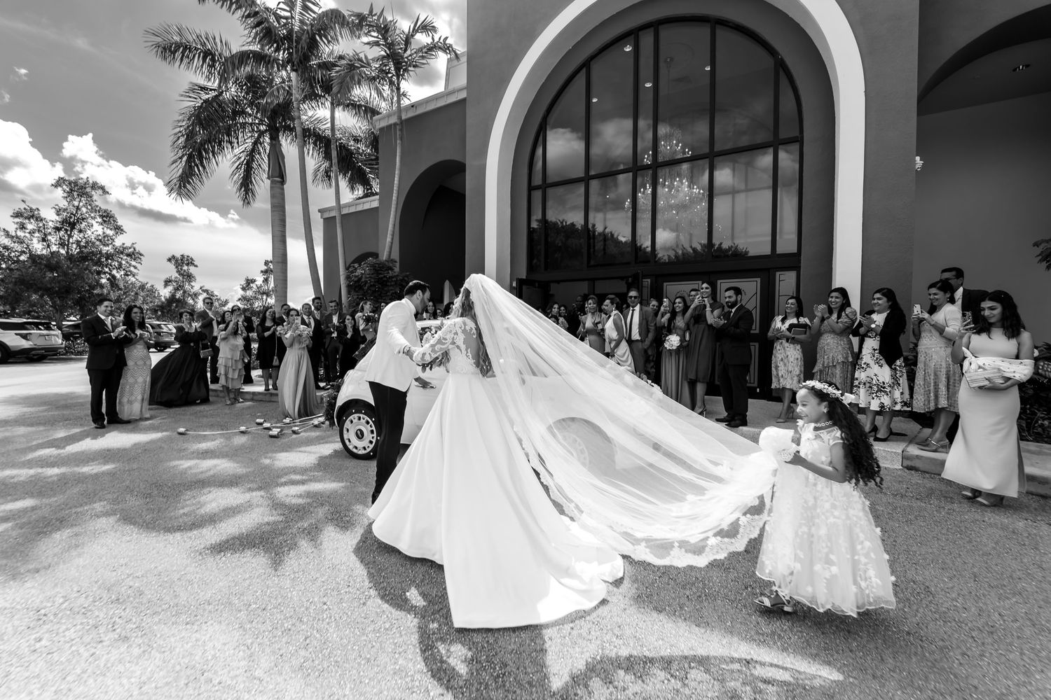 Foto B&W: Novia con velo largo baja de coche blanco ayudada por novio; invitados y niña flores esperan.