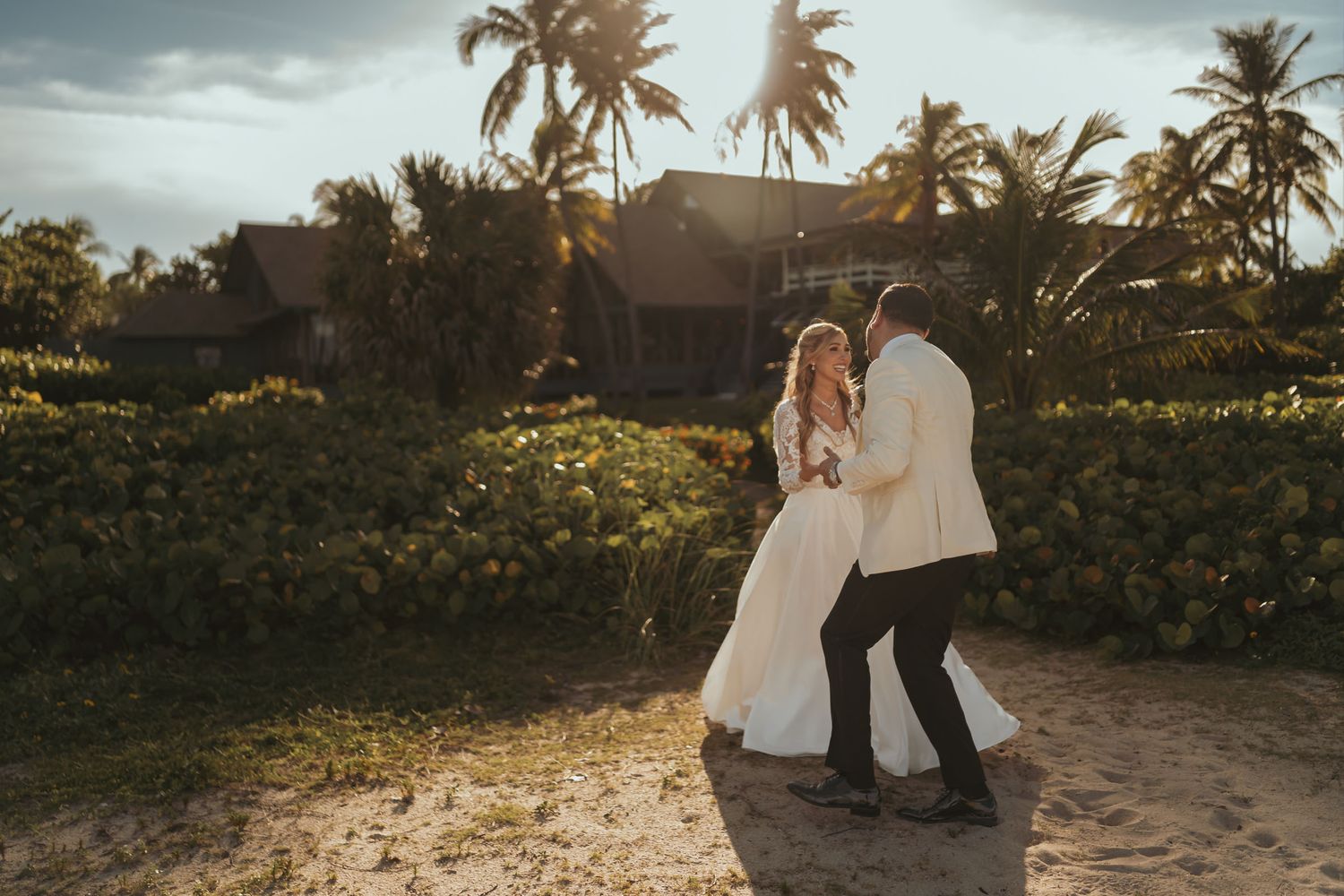Novios bailan o posan divertidos en playa con palmeras al atardecer; él chaqueta clara. Boda tropical.