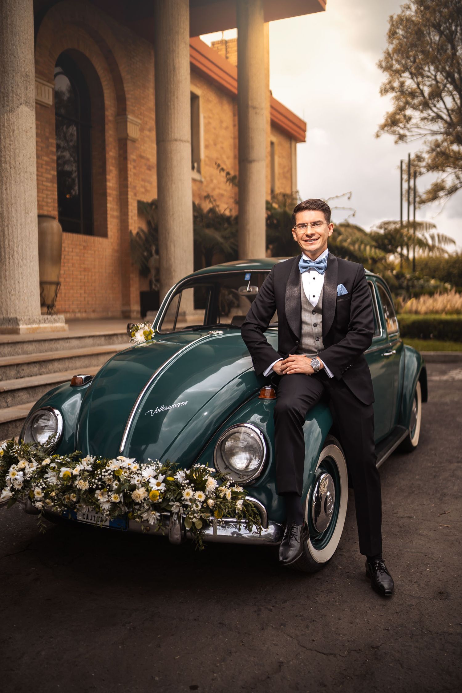 Novio con esmoquin posa sonriente sentado en capó de VW Escarabajo verde decorado para boda frente a edificio.