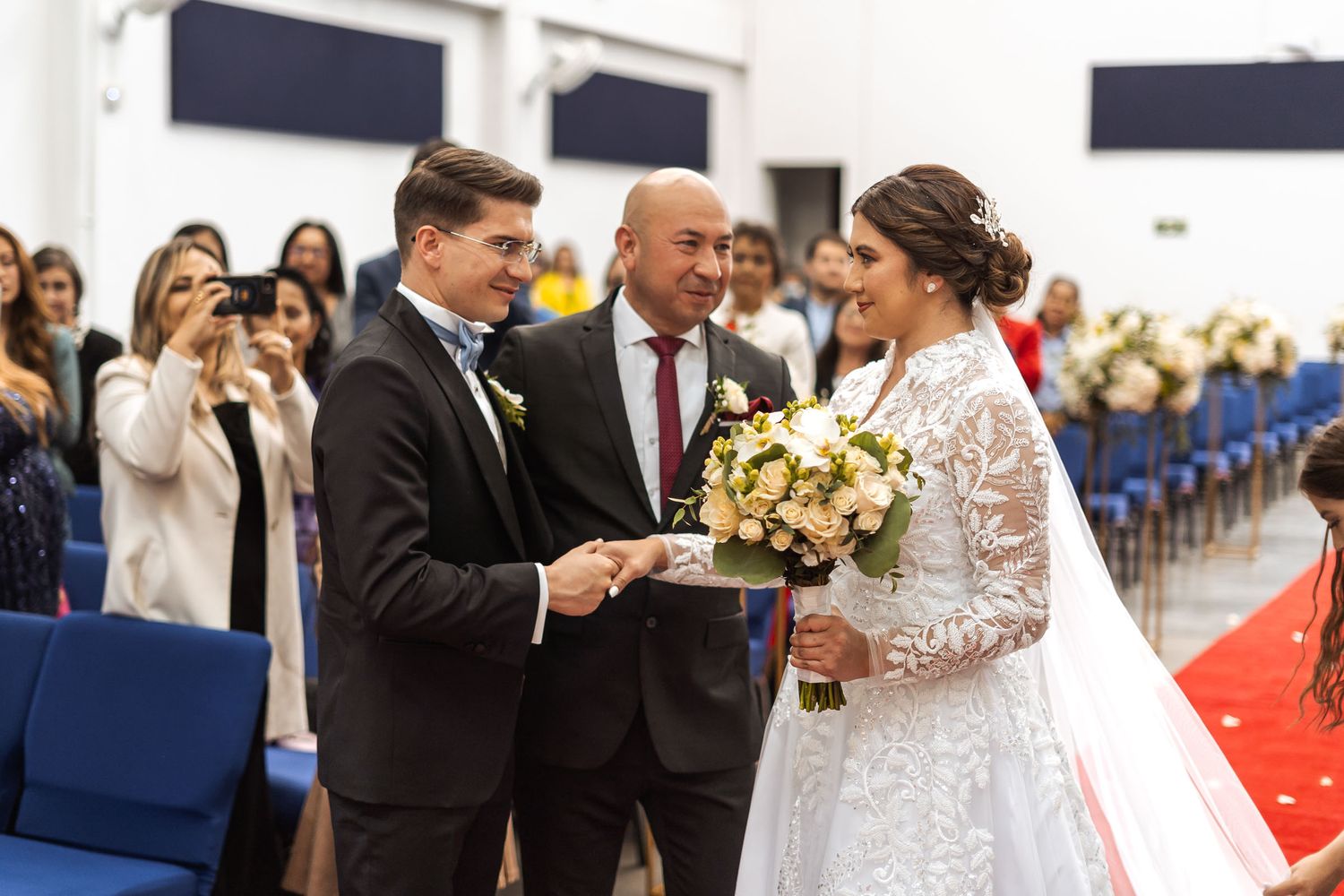 Padre entre la novia al novio en boda IDMJI. Invitados observan mientras ellos ingresan por el tapete rojo