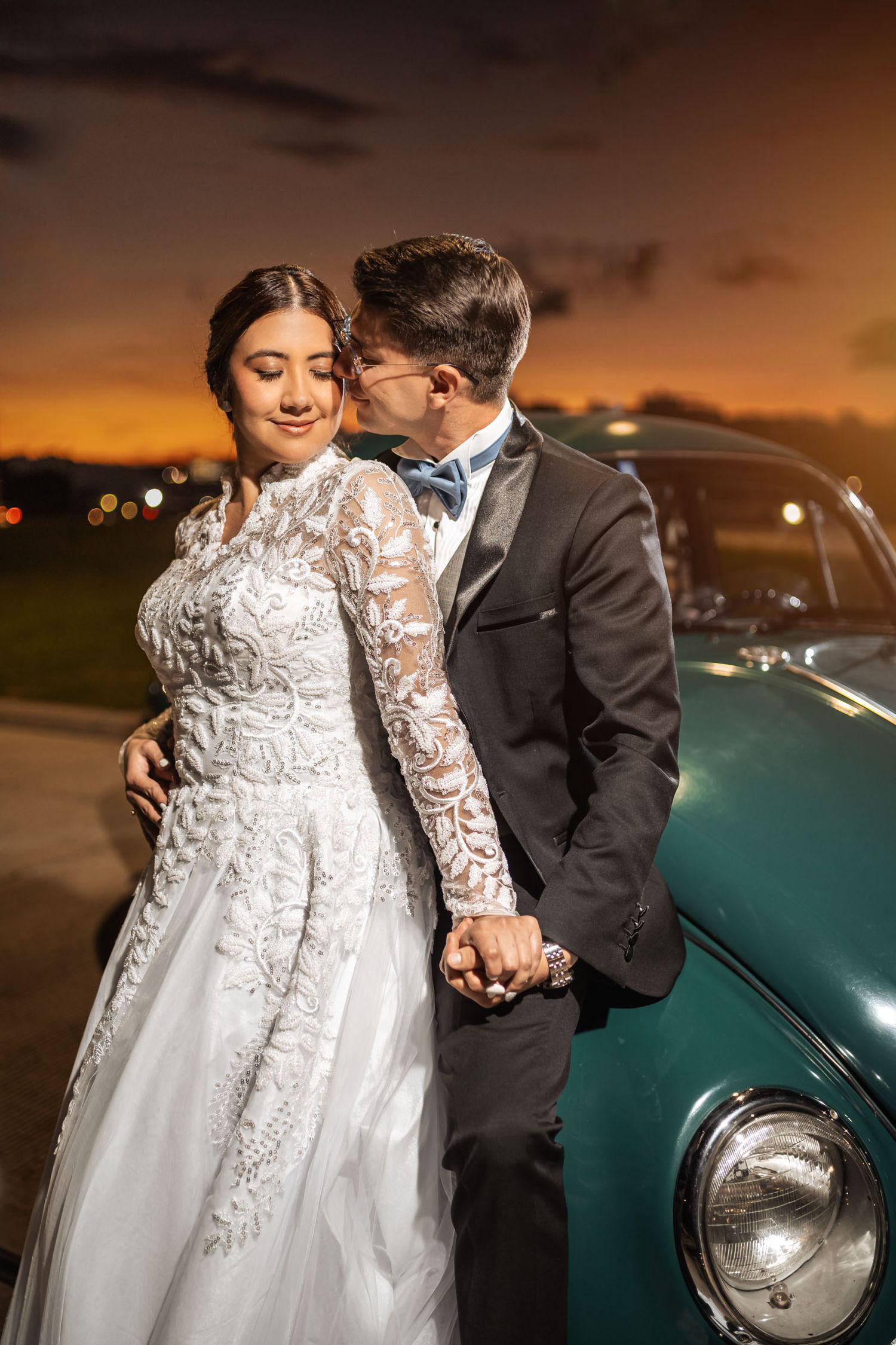 Novios tomados de la mano junto a coche verde clásico al atardecer. Él le susurra, ella sonríe. Boda romántica.