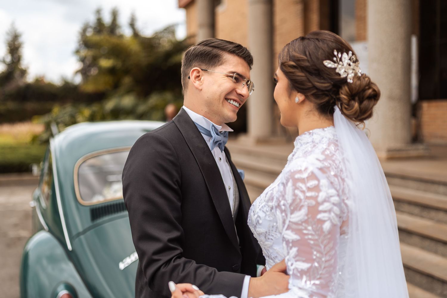 Novios sonrientes se miran frente a edificio con columnas; él con esmoquin, ella con vestido de encaje y velo. Coche clásico al lado.
