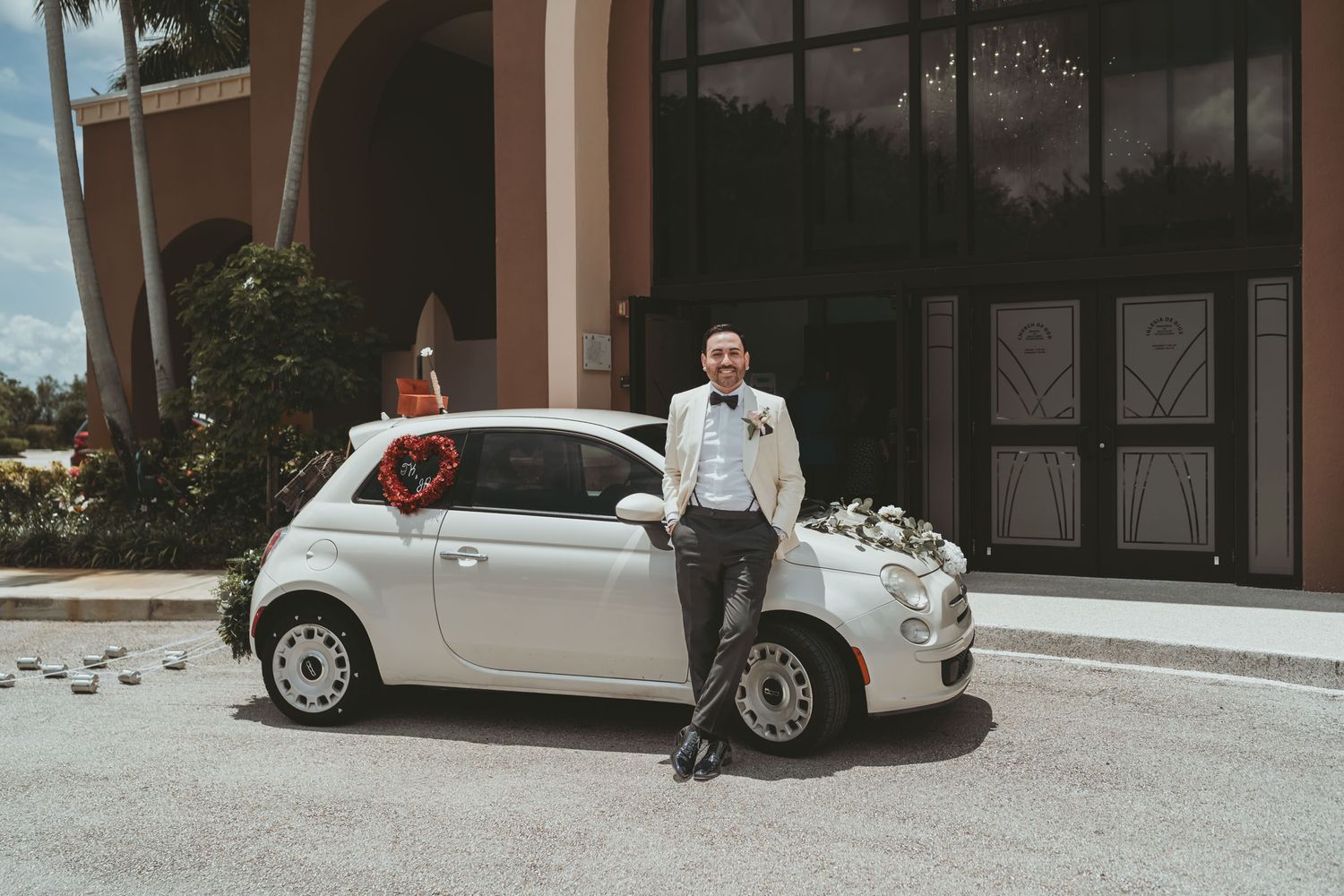 Novio elegante con traje blanco posa apoyado en coche pequeño blanco decorado para boda; exterior.