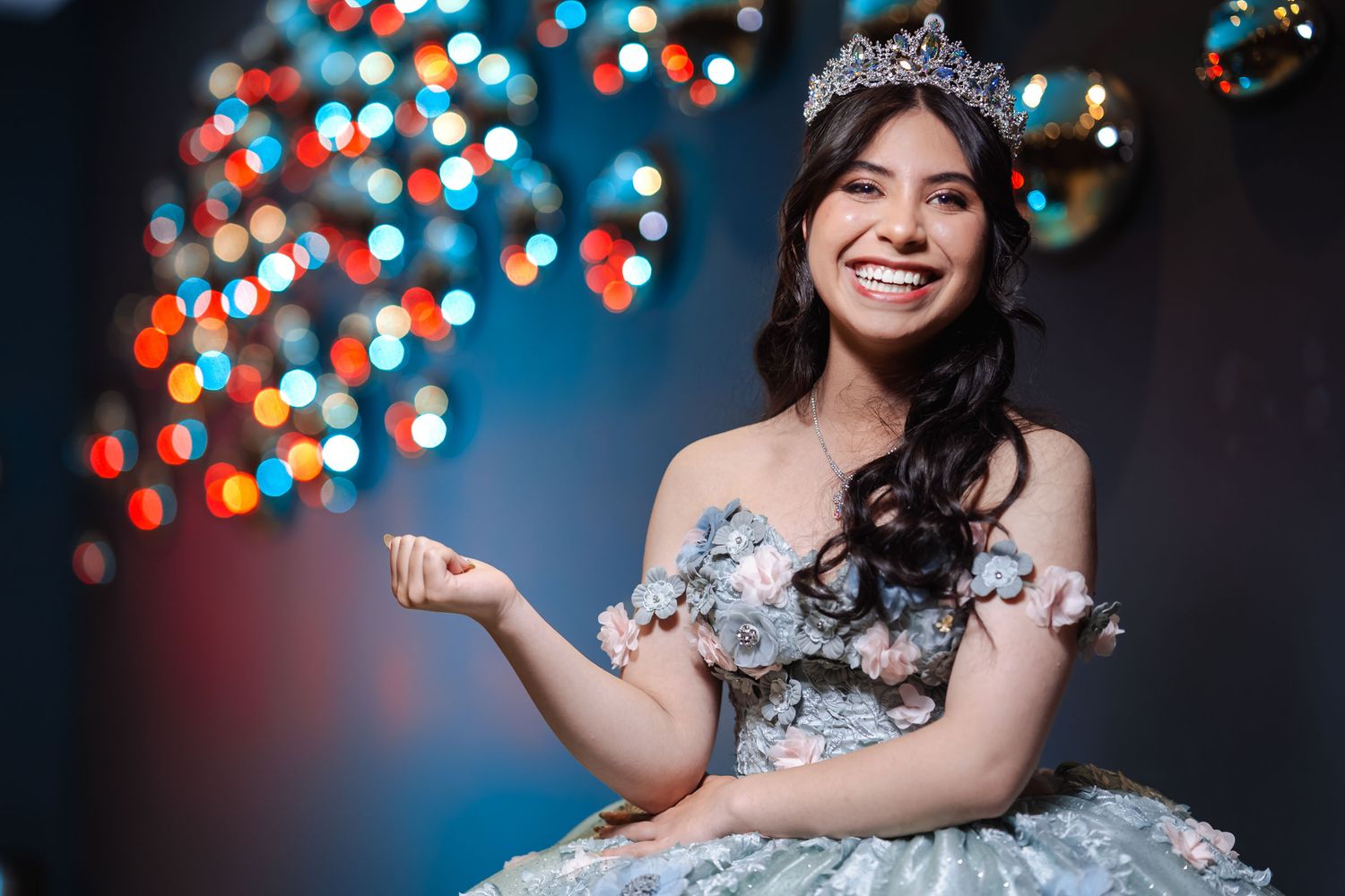 Retrato primer plano quinceañera radiante sonriendo; luce tiara y vestido bordado. Fondo luces bokeh coloridas.