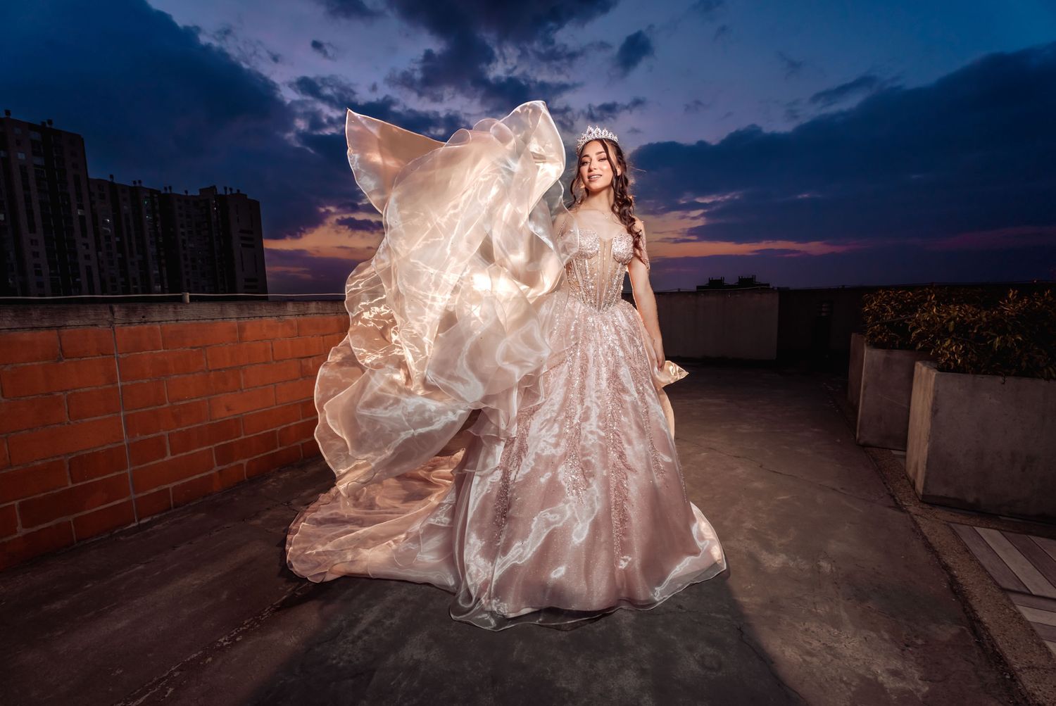 Quinceañera con vestido princesa rosa brillante y tiara en terraza; tela flota al atardecer, cielo dramático.