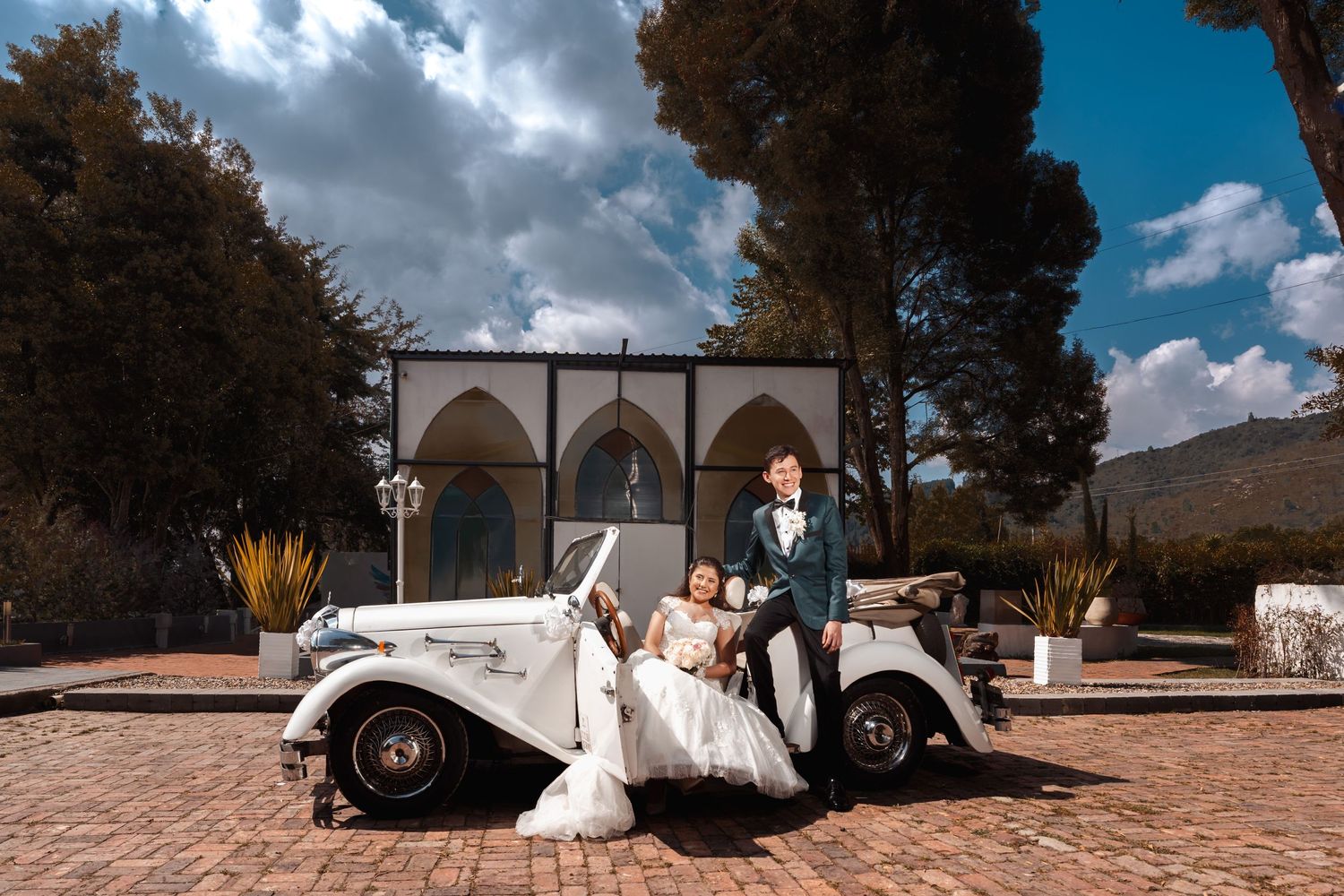 Elegante pareja de recién casados junto a coche clásico blanco en exterior y cielo hermoso. Novia con vestido blanco, novio con traje oscuro