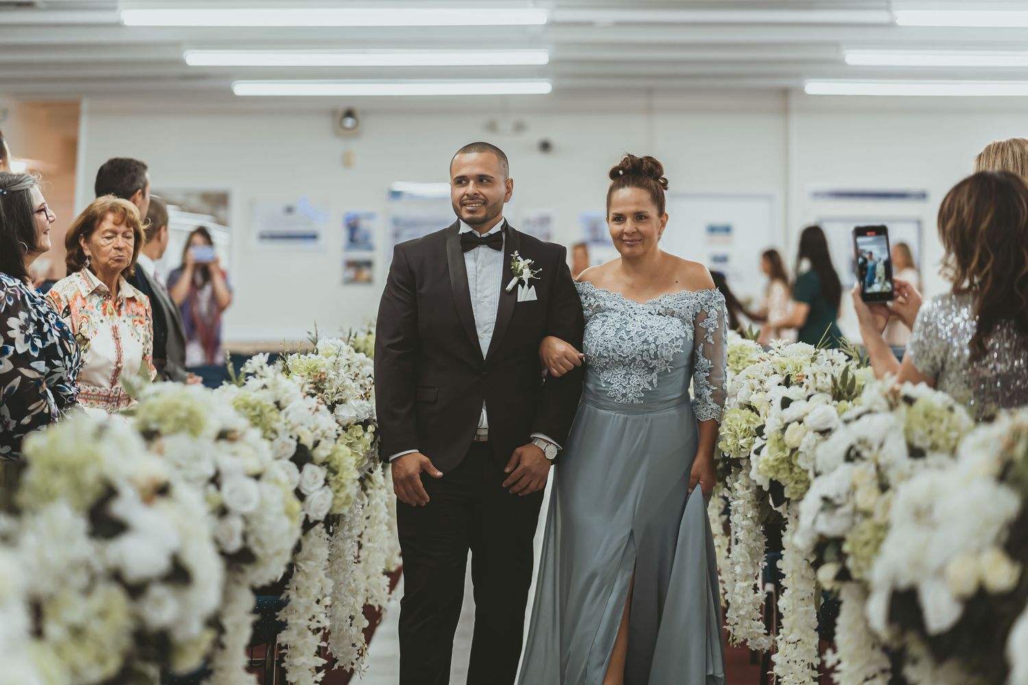 Novio camina del brazo de su madre (vestido azul/gris) por pasillo iglesia en entrada nupcial. Boda.
