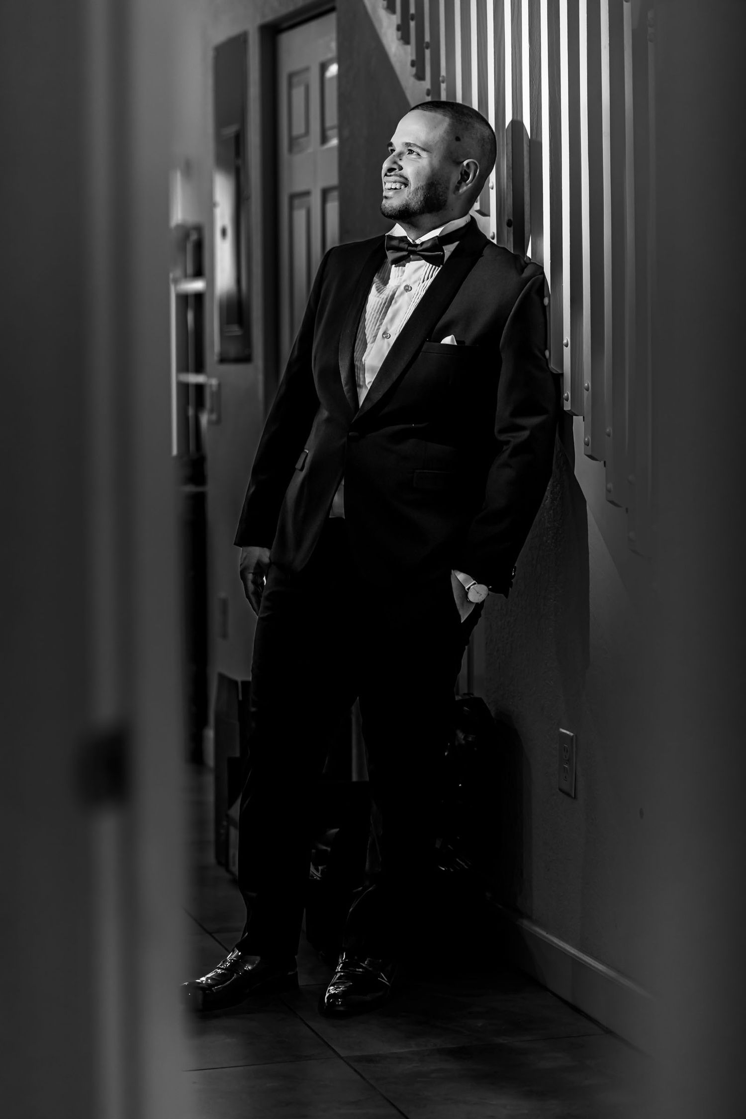 Foto B&W: Novio elegante con esmoquin apoyado en marco puerta interior, mira hacia luz. Retrato boda