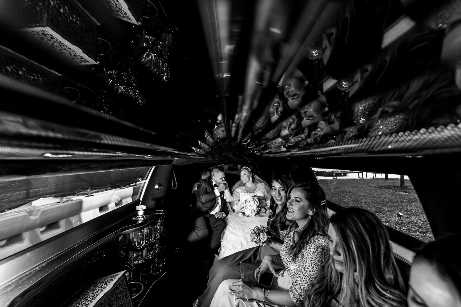 Foto B&W: Novios y cortejo nupcial en interior de limusina; techo reflectante, ambiente festivo. Boda.