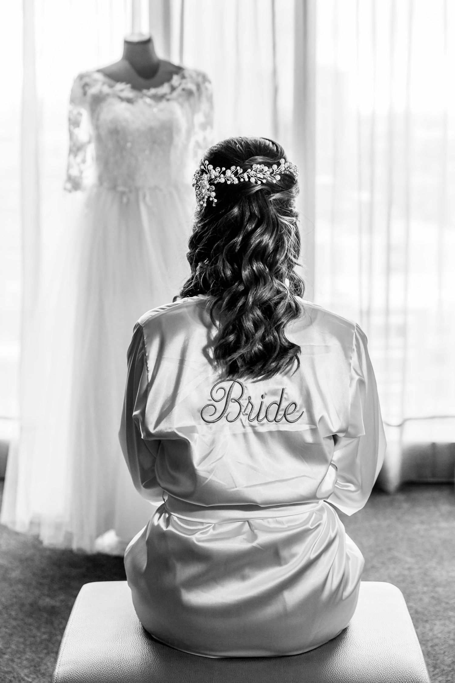 Foto B&W: Novia de espaldas con bata personalizada mira su vestido de novia colgado. Preparación boda.