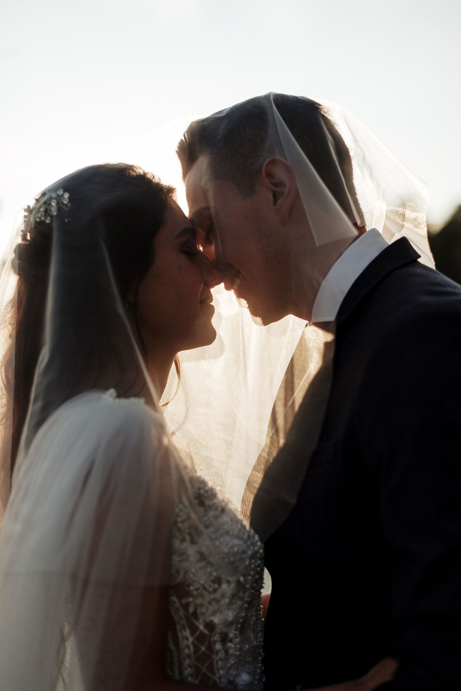 Primer plano de novios apunto de besarse a través del velo, con luz cálida. Momento íntimo de boda romántica.