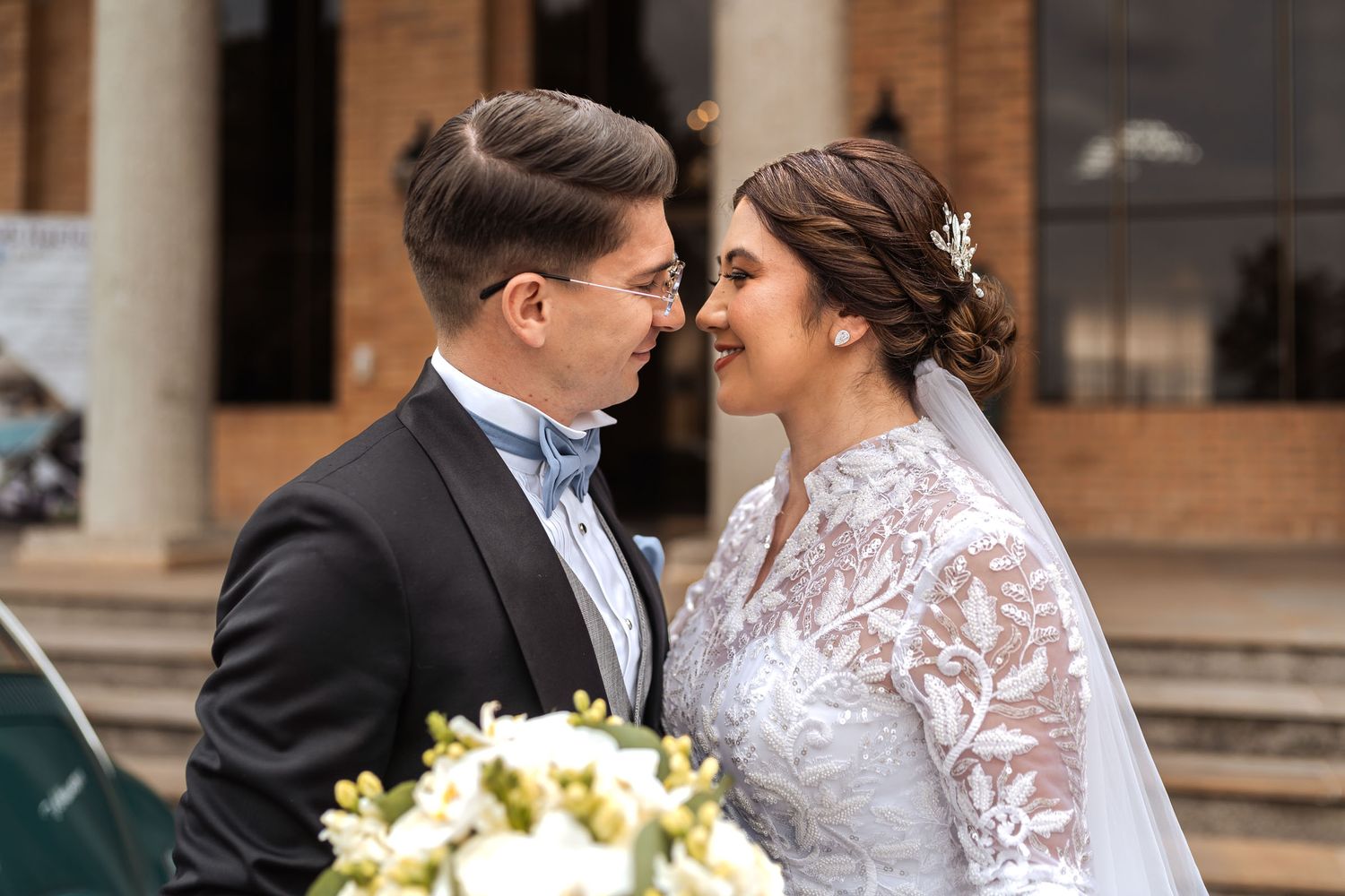 Novios sonrientes se miran frente a entrada de edificio; él con esmoquin, ella vestido de encaje y velo. Ramo de novia visible. Boda.