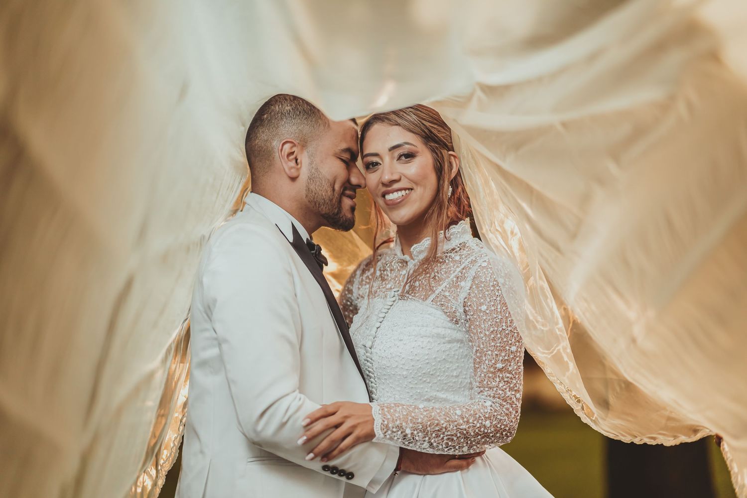 Retrato íntimo boda: Novios bajo velo o tela transparente; novia sonríe a cámara, luz cálida.