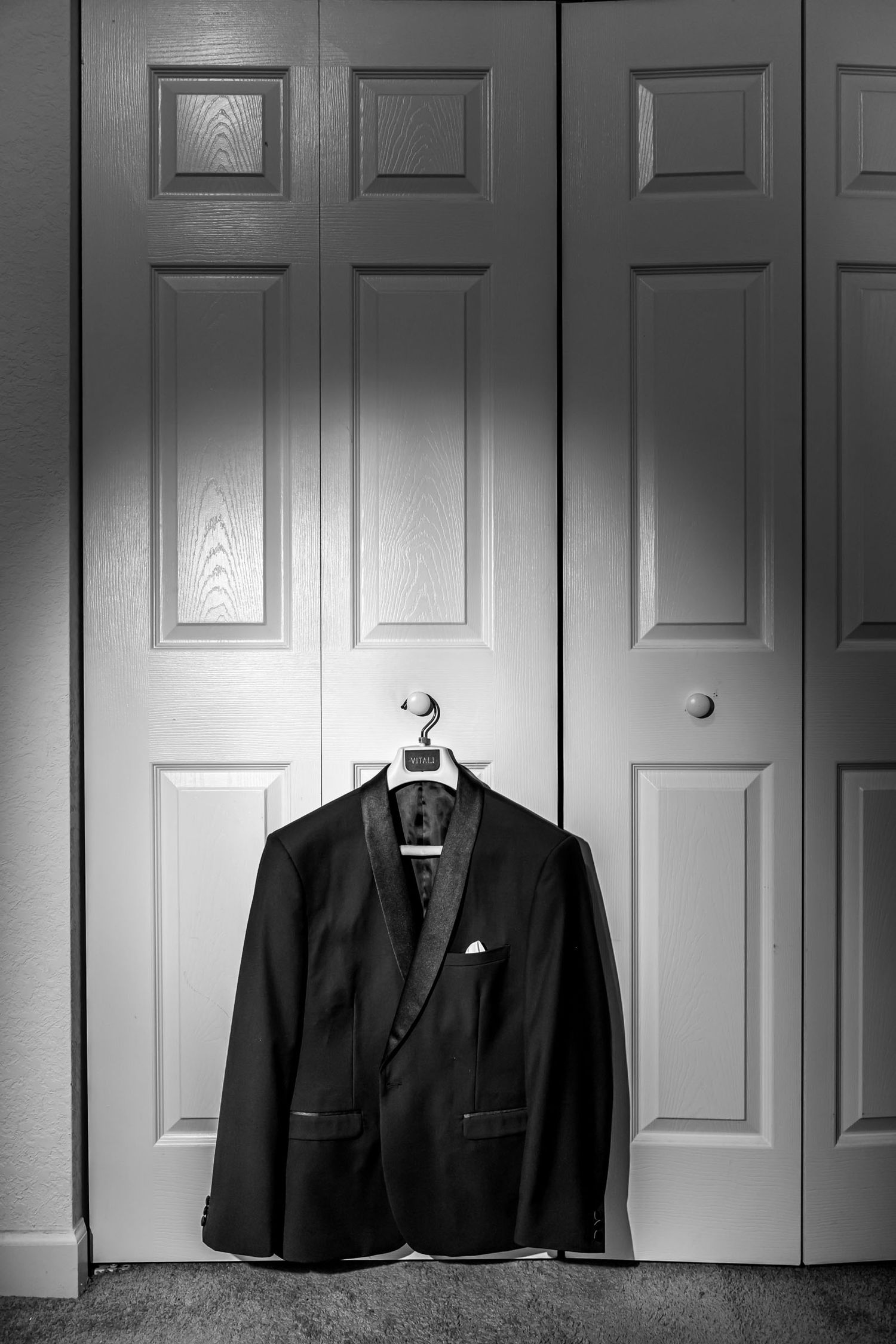 Foto B&W: Chaqueta esmoquin novio cuelga en puerta armario iluminada por luz cenital. Detalle boda.
