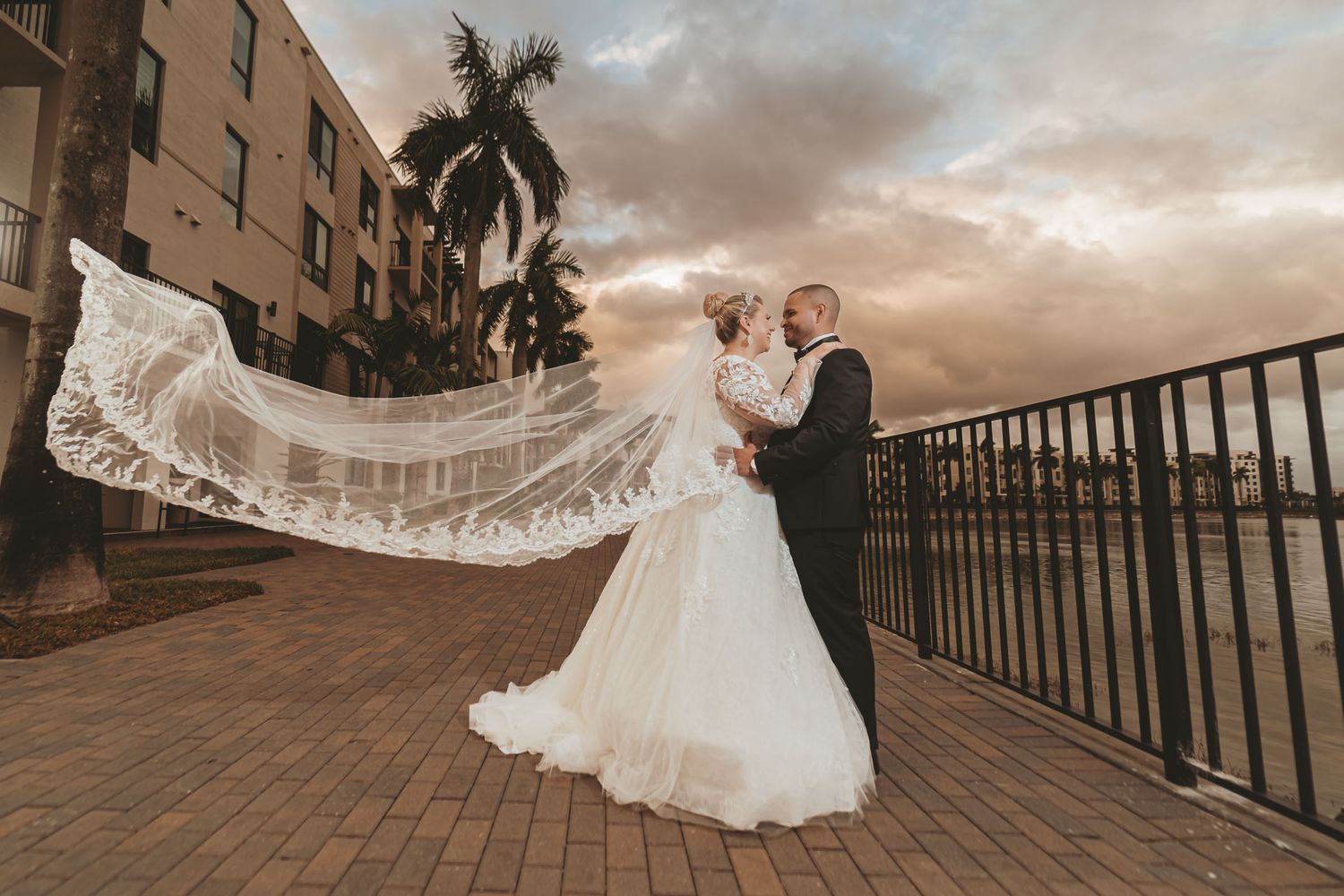 Novios posan en terraza al atardecer; velo largo de encaje de la novia ondea espectacularmente al viento. Boda en Miami.