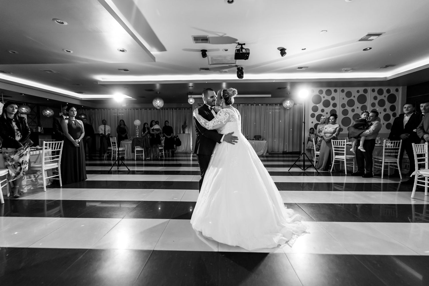 Foto B&W: Novios comparten primer baile en pista a cuadros blanco y negro durante recepción de boda; invitados miran.