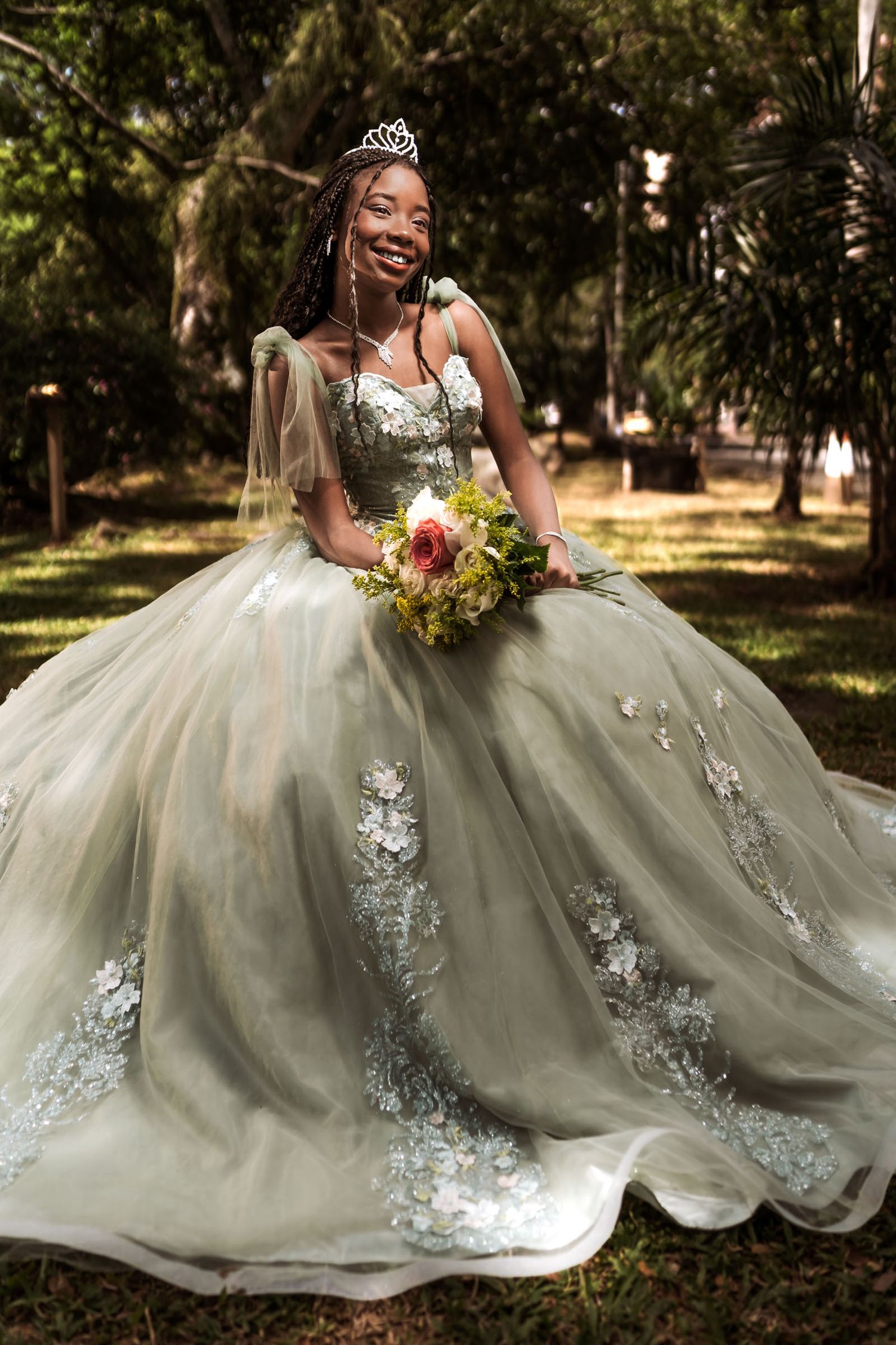 Quinceañera afrodescendiente (trenzas) sonríe sentada en césped con vestido princesa verde/plata y tiara.