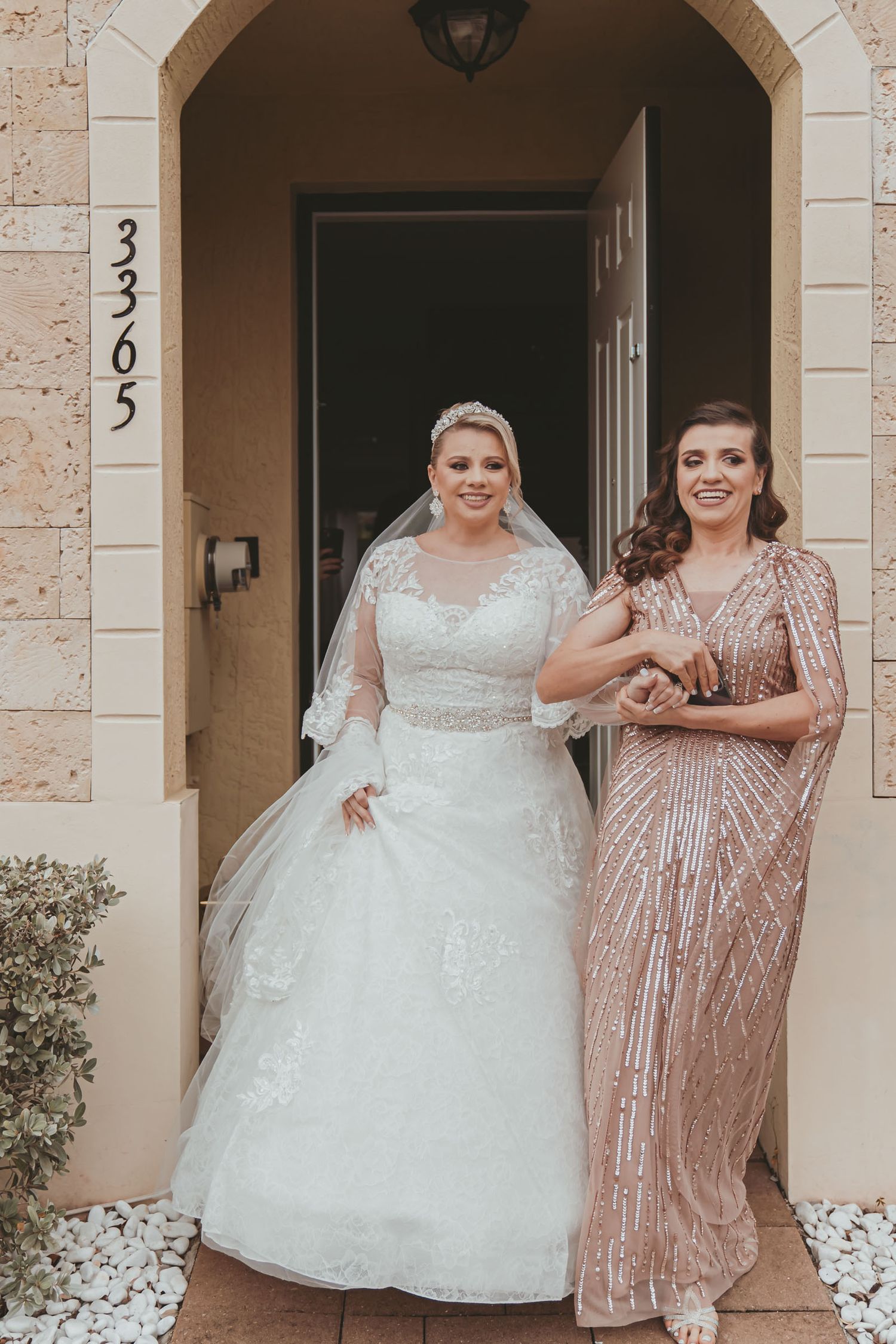 Novia sonriente con vestido y velo junto a su hermana (vestido brillante) en puerta de casa; preparación boda Miami.