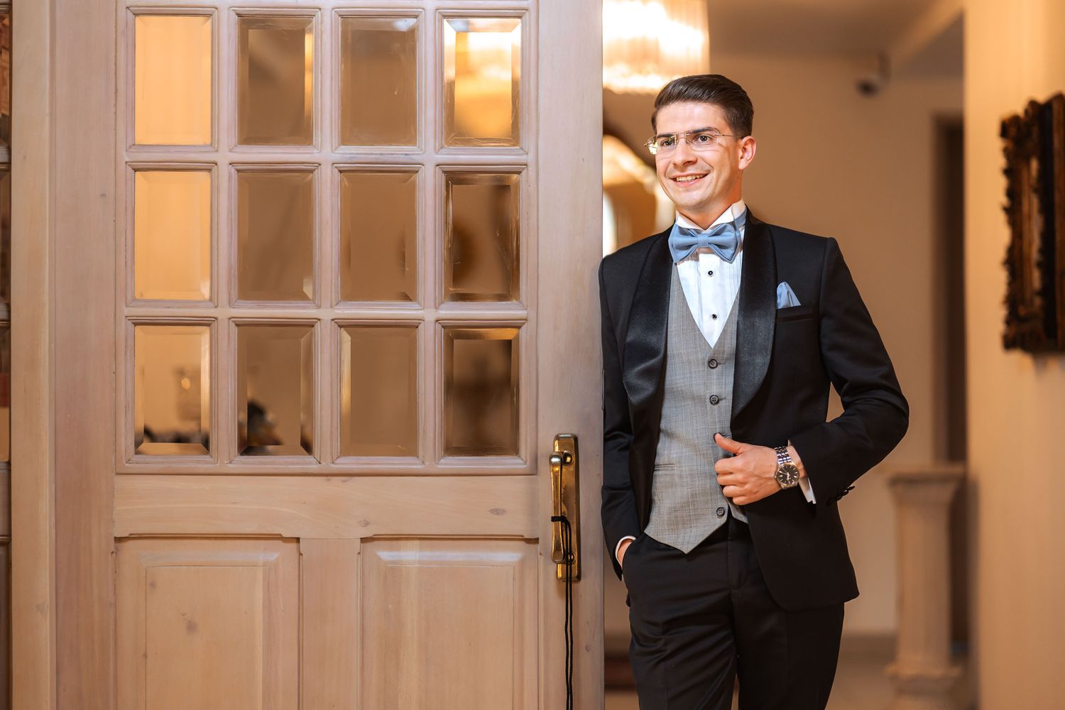 Novio con esmoquin posa relajado apoyado en puerta de madera con cristales en interior elegante; fotografía de boda.