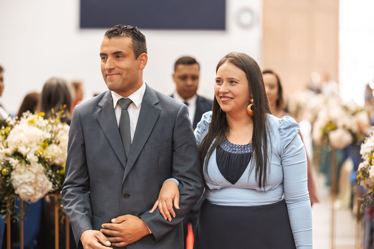 Pareja sonriente camina por pasillo de iglesia durante procesión de boda; él con traje gris, ella vestido azul.