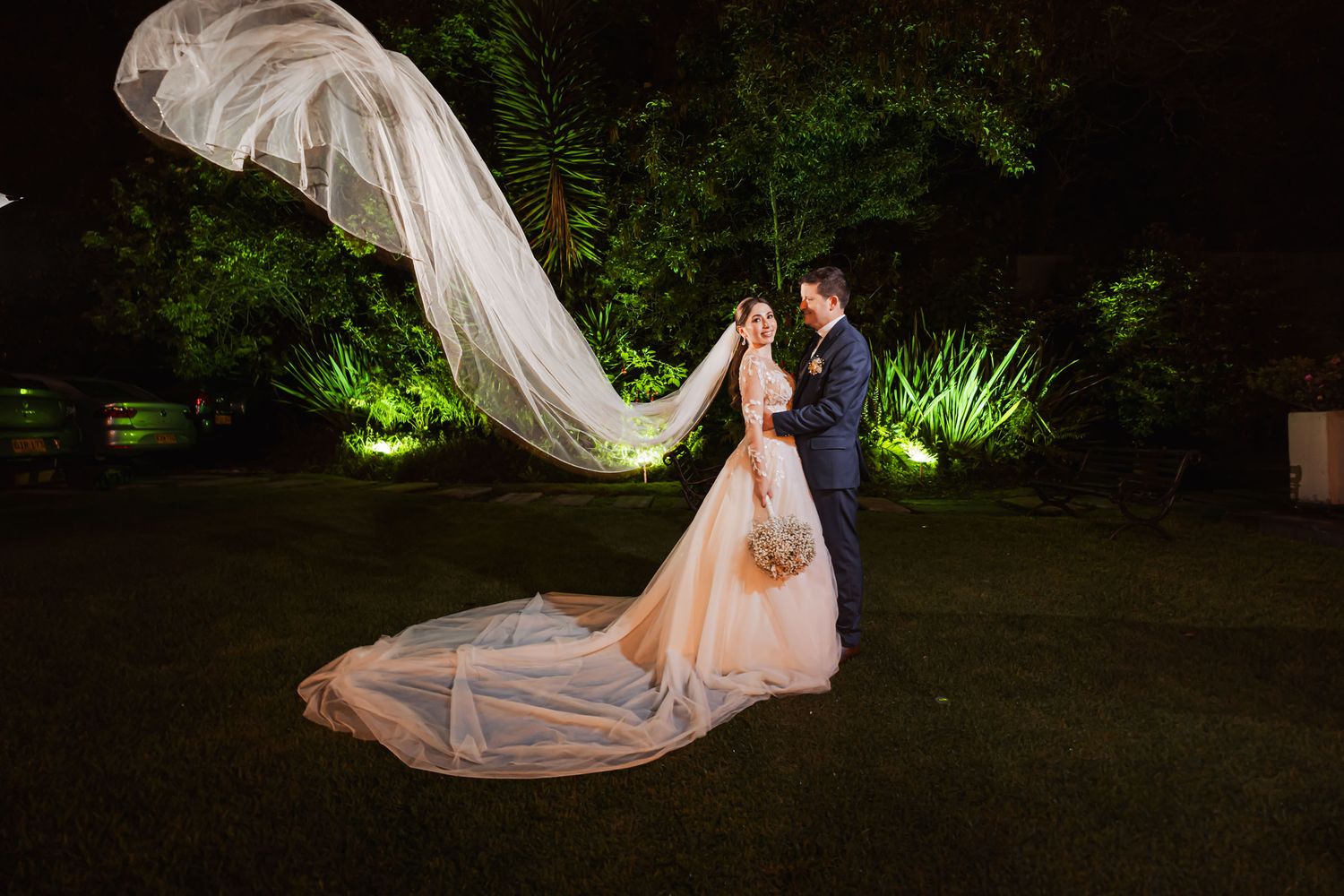 Novios posan de noche en jardín; velo largo de novia flota espectacularmente en el aire. Él traje azul. Boda.