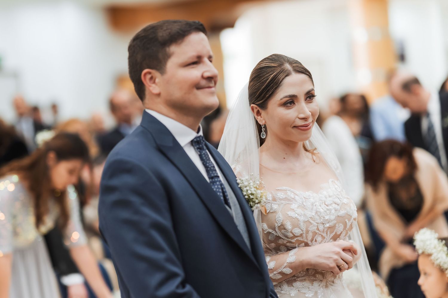 Novios sonrientes tomados de la mano durante ceremonia de boda en iglesia; invitados y niña de flores visibles.