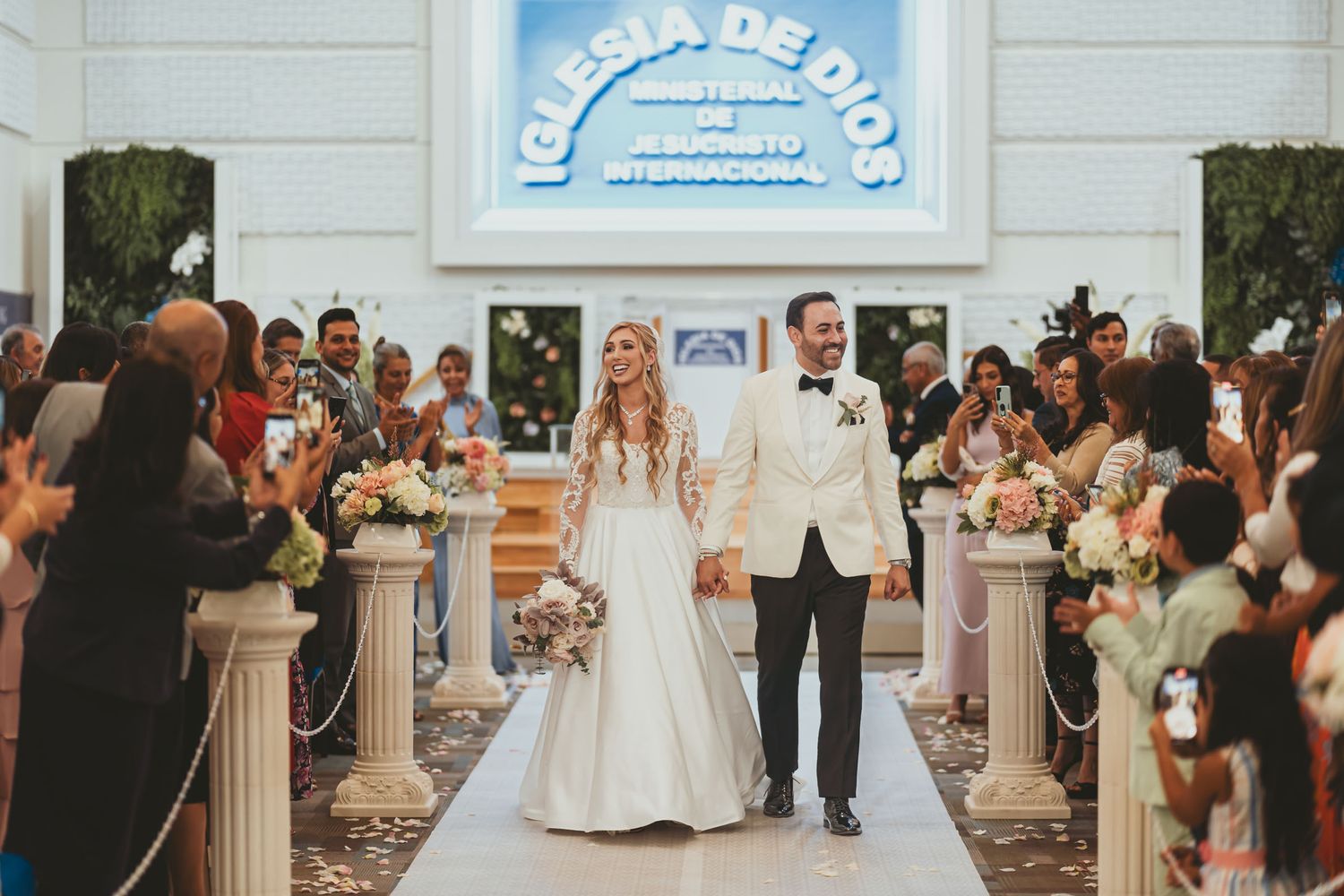 Salida nupcial: Novios caminan por pasillo iglesia (Iglesia de Dios); invitados toman fotos y aplauden. Boda IDMJI