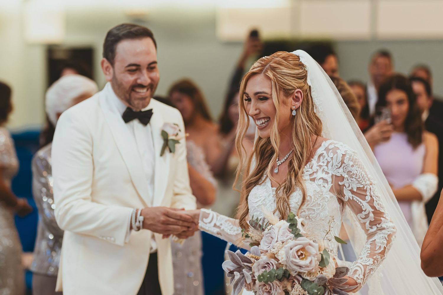 Novios sonrientes tomados de la mano en altar durante ceremonia boda; ella con ramo, él traje blanco.