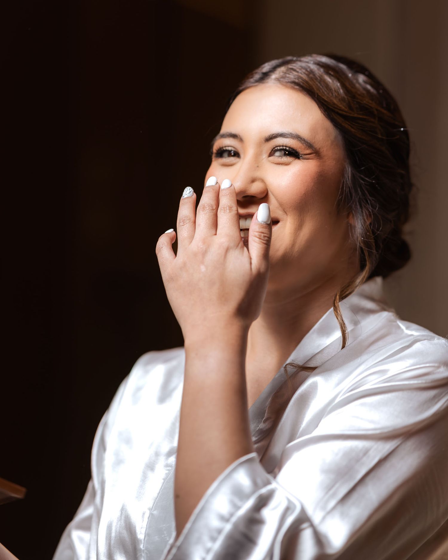 Novia ríe feliz cubriendo su boca con la mano (manicura blanca) en bata de satín; preparación de boda.