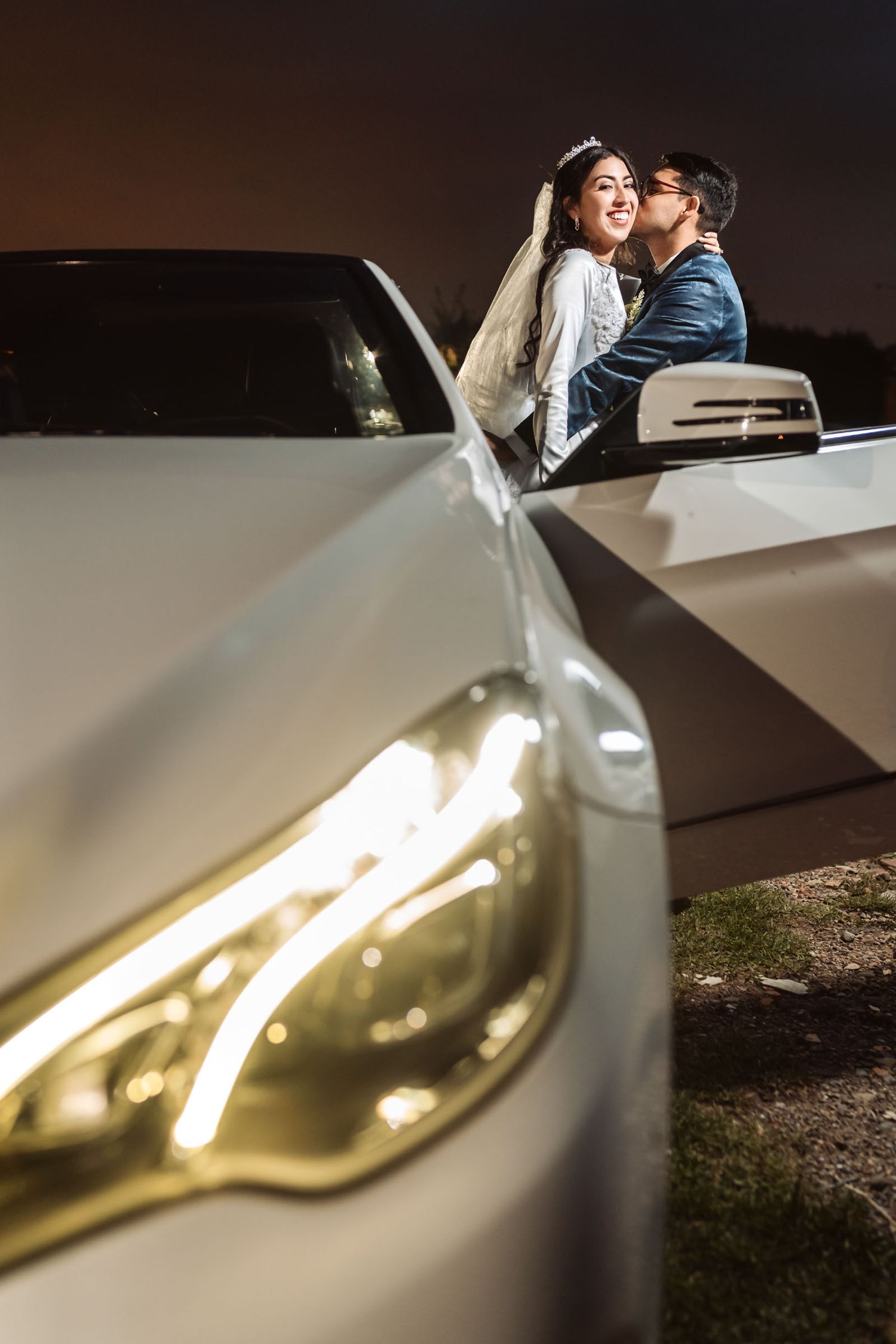 Novios se besan sobre capó de coche blanco de noche; faro encendido. Momento romántico boda.