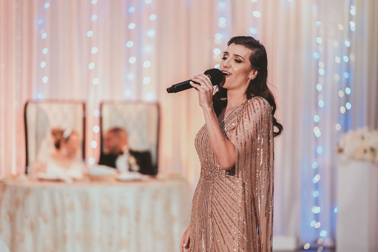 Cantante elegante con vestido brillante actúa con micrófono en recepción boda; novios fondo desenfocado.