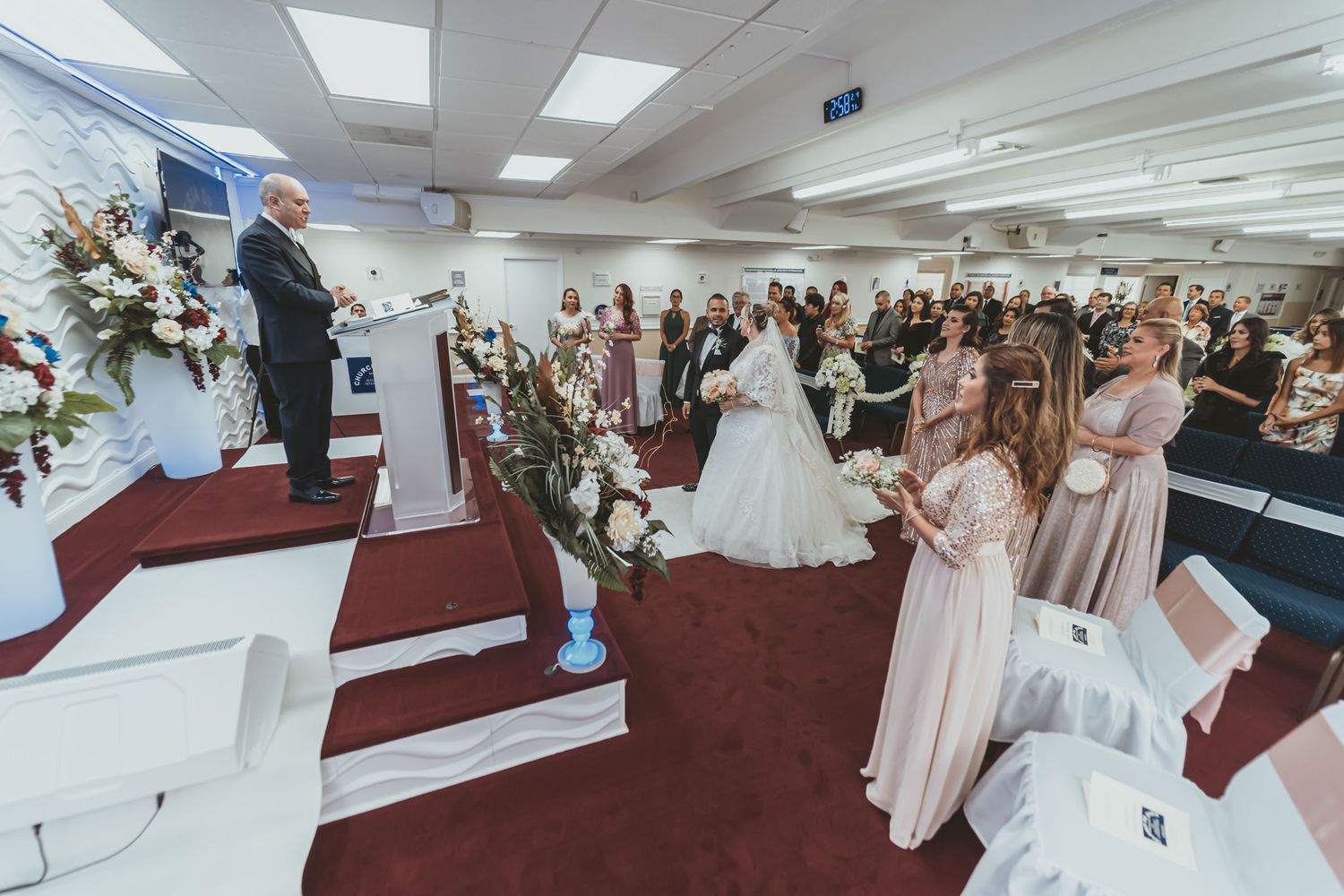 Vista lateral ceremonia boda: Oficiante en púlpito, novios e invitados de pie en iglesia.