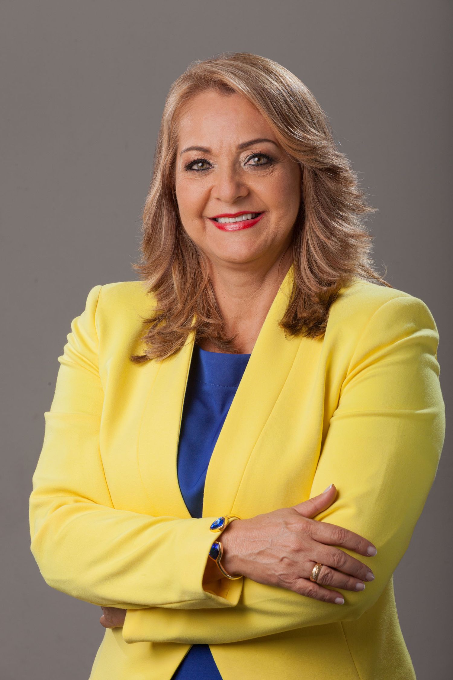 Retrato headshot mujer sonriente con blazer amarillo vibrante y top azul, brazos cruzados; fondo gris estudio. Aydeé Lizarazo