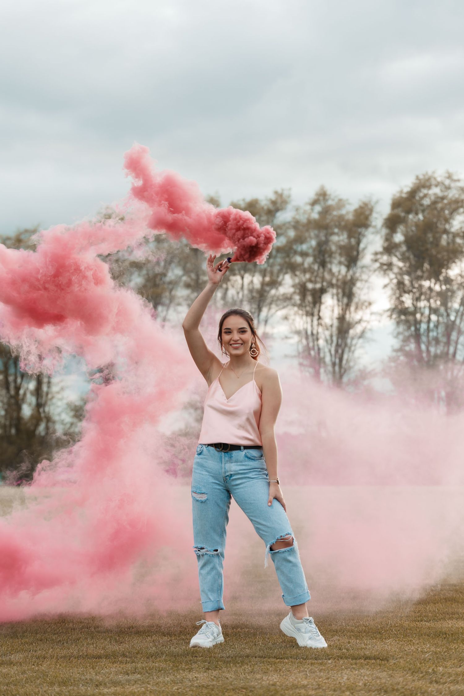 Mujer joven con top rosa y jeans rotos sostiene bengala de humo rosa en campo; cielo nublado. Sesión fotos prequince.
