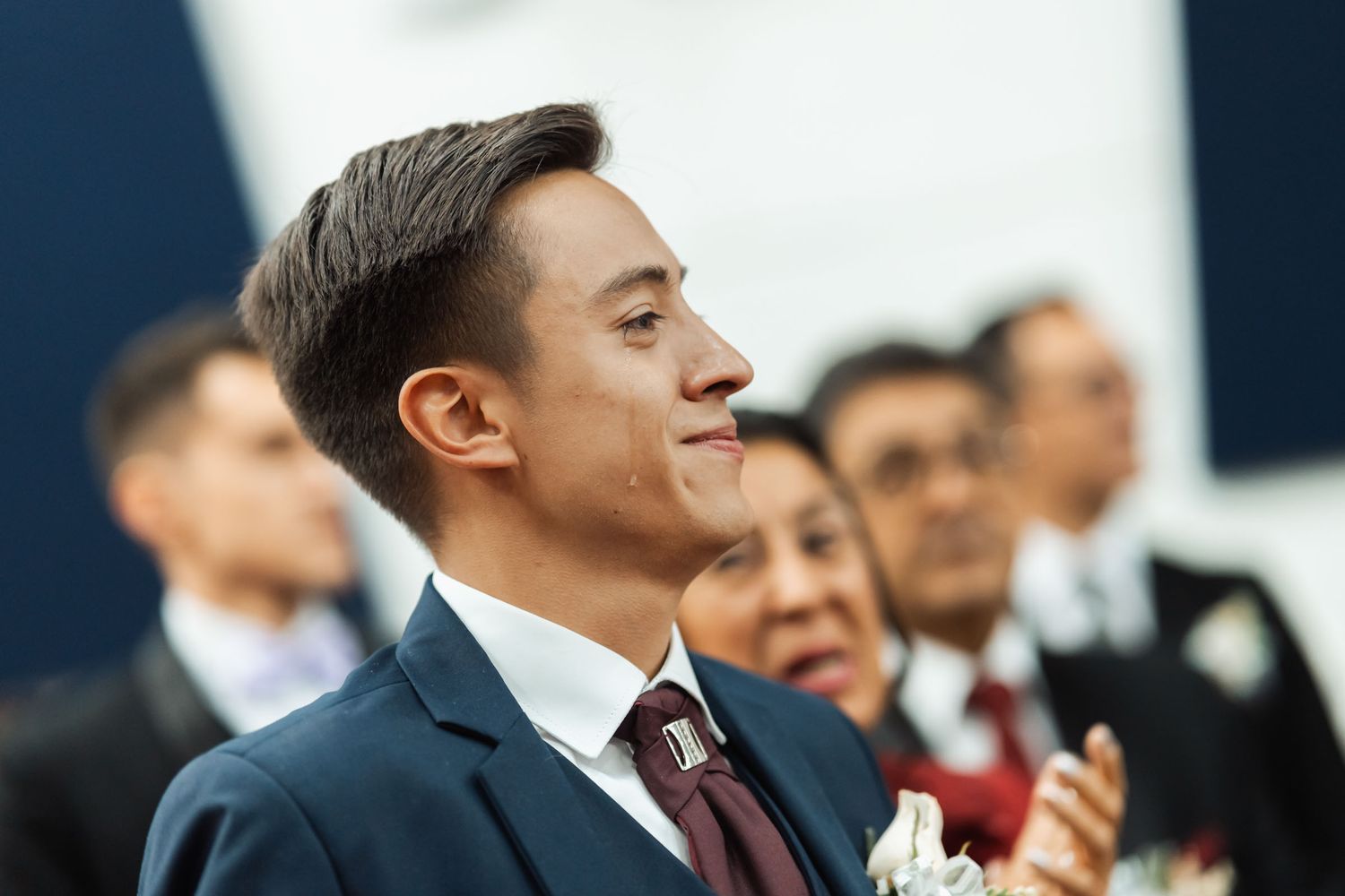 Novio con lágrima en mejilla mira conmovido durante ceremonia de boda; traje azul oscuro, corbata borgoña.