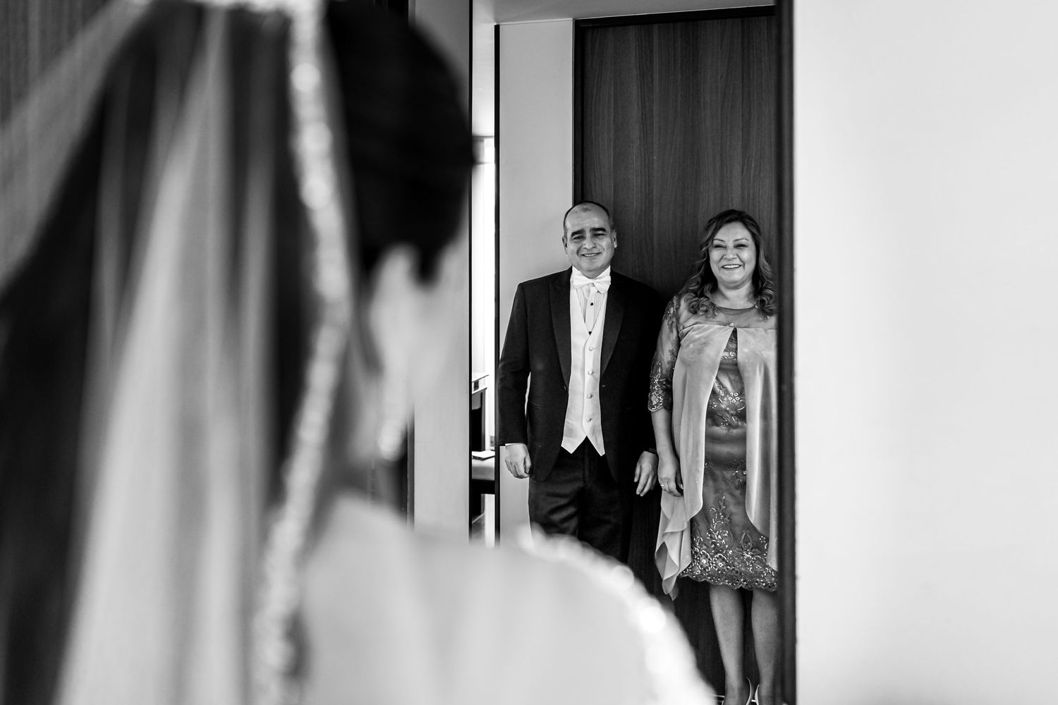Foto B&W: Padres miran conmovidos a novia (vista espalda) al verla vestida; first look. Alistamiento novia boda