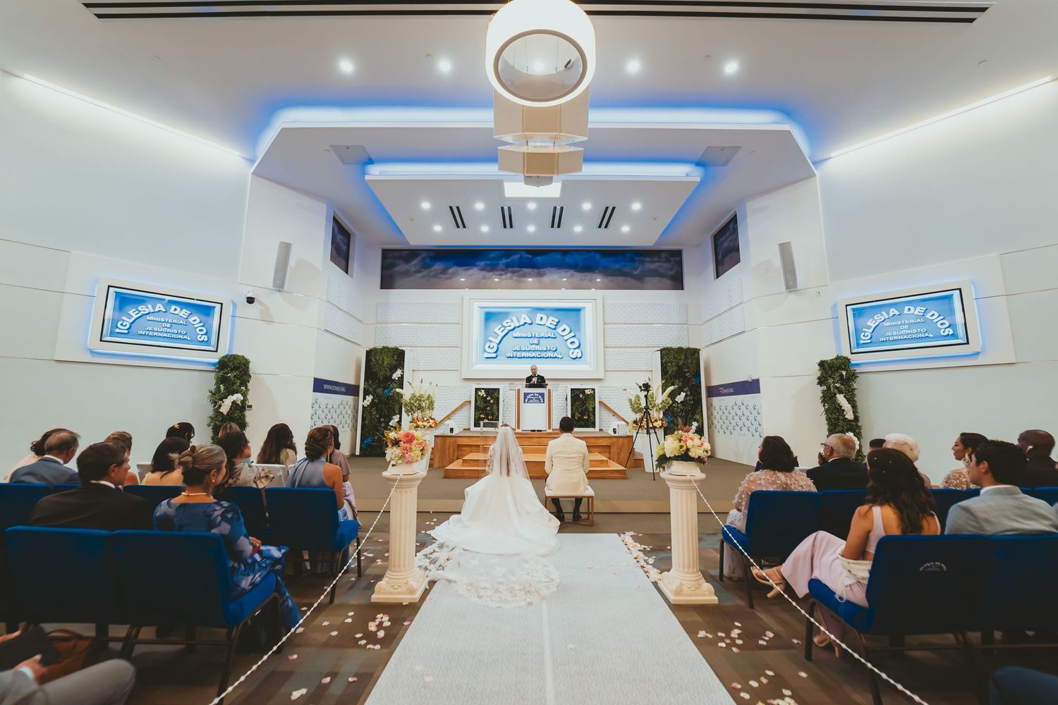 Vista amplia desde atrás de iglesia/salón moderno durante ceremonia boda; novios en altar, invitados.
