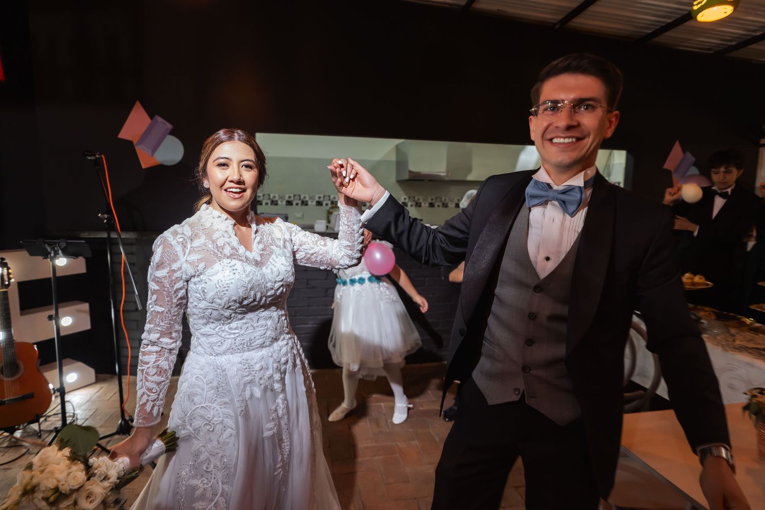 Novios bailan felices en la recepción de boda, tomados de la mano. Una niña aparece detrás. Ambiente festivo.